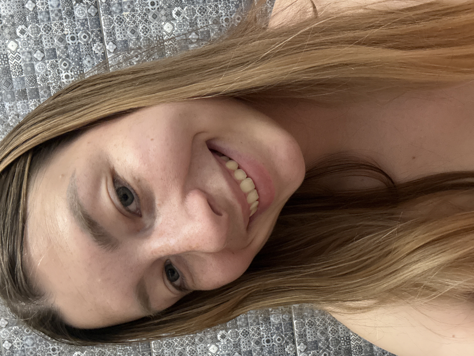 A close-up, angled selfie of a woman with long, light brown hair. She is smiling, revealing her teeth. Her eyes are blue-grey. The background appears to be a patterned tile surface. There is no evidence indicating the location is Moscow, Russia.