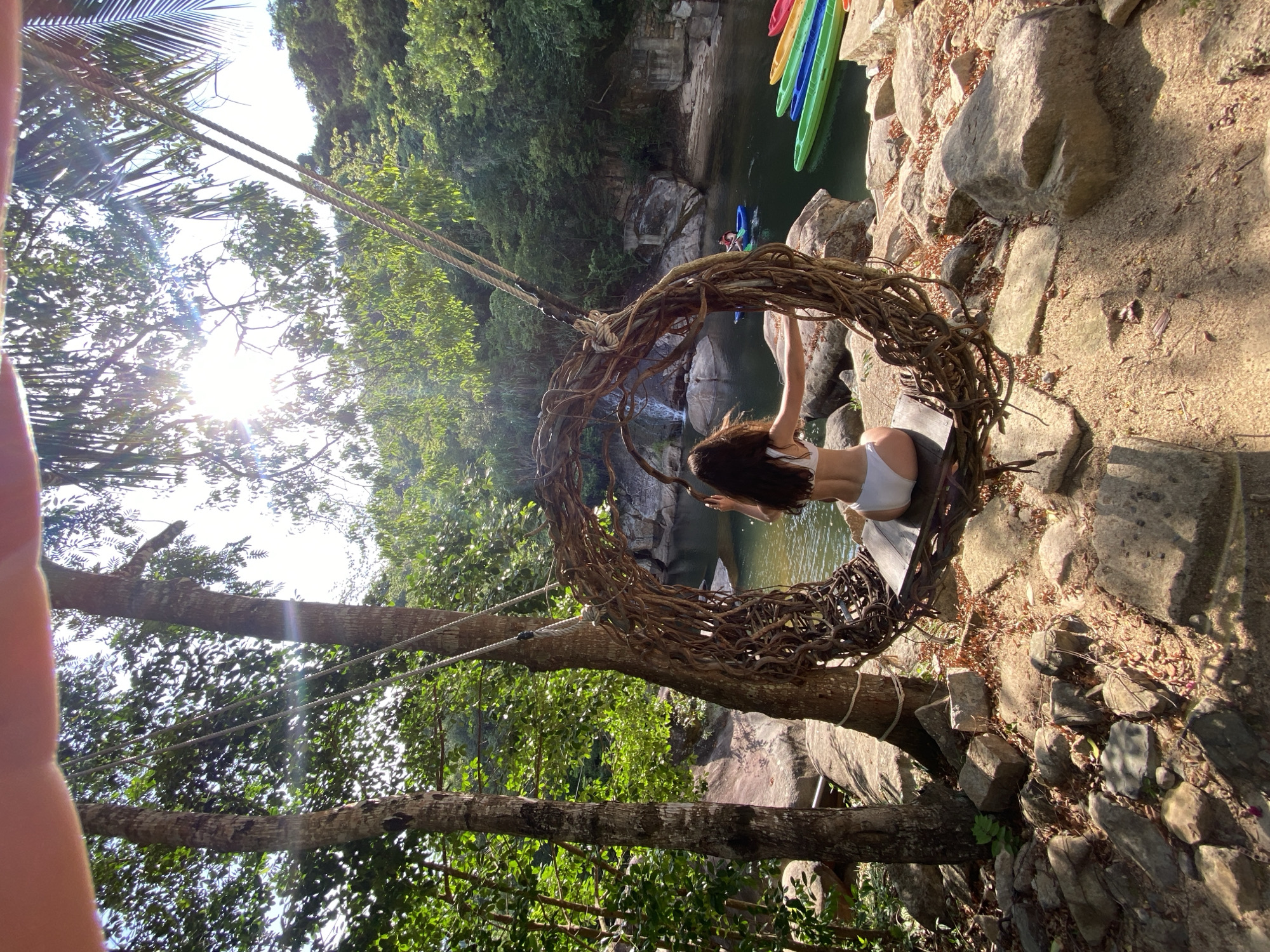 A woman wearing a white bikini is sitting on a wooden swing with a circular frame made of branches. The swing is suspended between two trees. She is facing away from the camera, looking towards a body of water. In the water, there are several kayaks of different colors lined up. Further in the distance, there is a small waterfall. The environment is lush and green, with trees and rocks surrounding the area. The sun is shining brightly, casting shadows and creating lens flares in the image. The scene appears to be in a natural, outdoor setting, likely a tourist attraction or resort in Hoa Thang Ward, Vietnam.