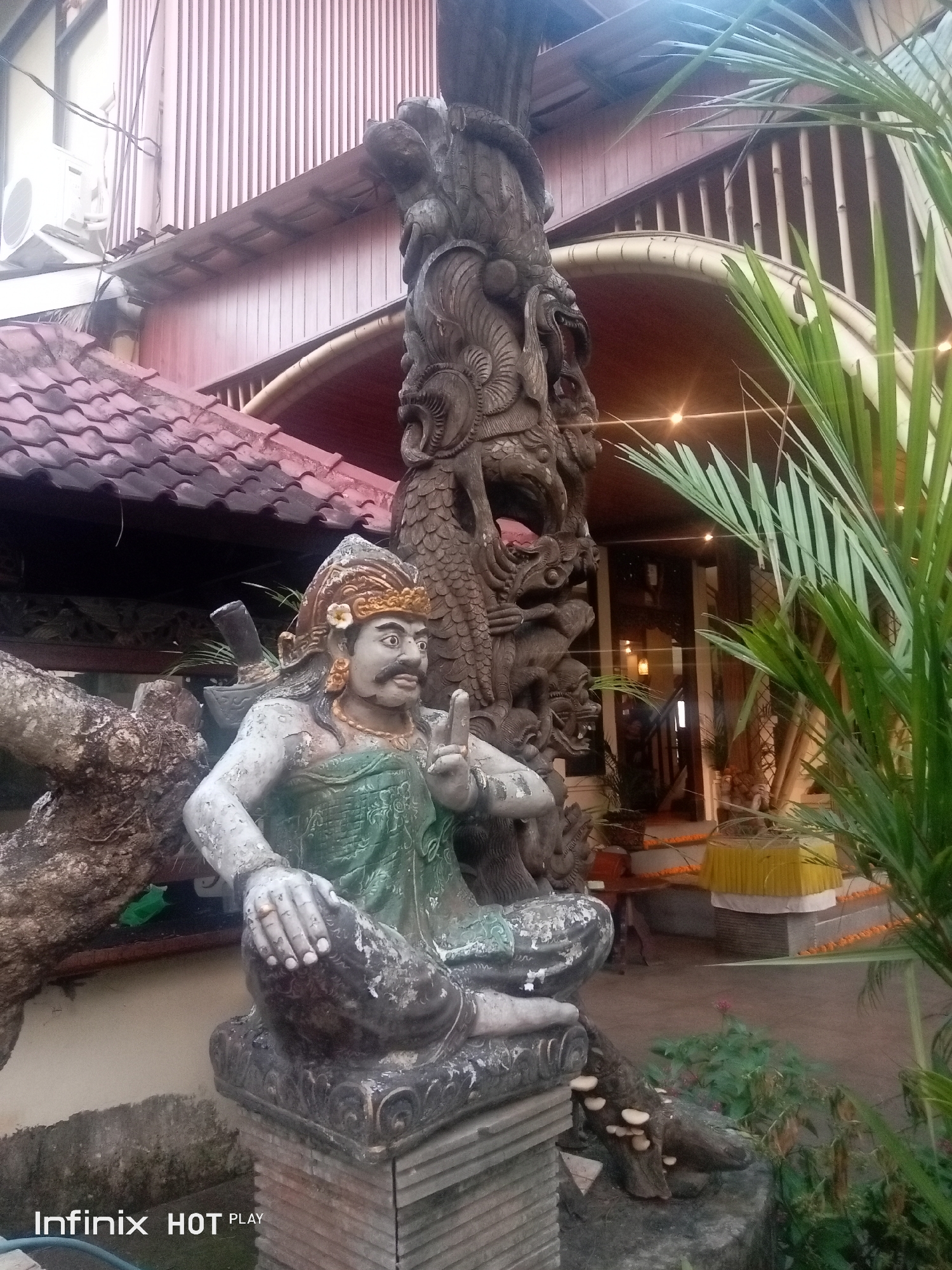 In the foreground of the image, a weathered, stone statue of a figure is seated on a tiered platform. The figure wears a golden headdress with a white flower tucked into the side, a necklace, and a green garment. Its skin is a pale, chipped grey. The figure's right hand is raised, with two fingers extended in a gesture. To the left of the statue, a gnarled, tree-like form is visible, also appearing to be made of stone or aged material. Behind the statue, a large, intricately carved wooden totem pole stands, depicting mythological figures and serpentine forms. To the right, lush green palm fronds partially obscure the background.
In the midground, the entrance to a building is visible. The architecture features a sloped, tiled roof over an open-air area, supported by wooden beams and a curved structure. Vertical wooden slats form a wall or screen behind this. An air conditioning unit is mounted on the wall of the building to the left. The interior of the building, visible through an opening, shows a staircase and dimly lit spaces. Yellow and orange decorative elements are placed on the ground near the entrance.
The overall environment suggests an outdoor setting at a cultural or religious site, possibly a temple complex or a garden in a resort, within the Ubud District of Indonesia. The lighting indicates it might be late afternoon or early evening, with artificial lights starting to illuminate the interior.