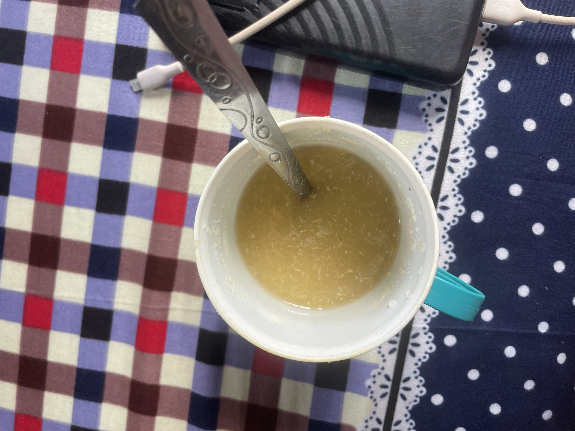 The image shows a close-up, overhead view of a white mug filled with a light-colored, textured liquid, possibly a soup or porridge. A patterned spoon is partially submerged in the liquid. The mug has a teal-colored handle. The mug and spoon are placed on a surface covered by a checkered cloth with alternating squares of blue, purple, red, brown, and white. Below this checkered cloth, a blue cloth with white polka dots is visible, edged with a white lace trim. To the right of the mug, a black object, possibly a phone or power bank, is visible, with a white charging cable plugged into it. The overall setting appears to be indoors. Without further information or visual cues, it is not possible to determine the specific location within Keffi, Nigeria, or to infer any details about people, actions, or interactions.