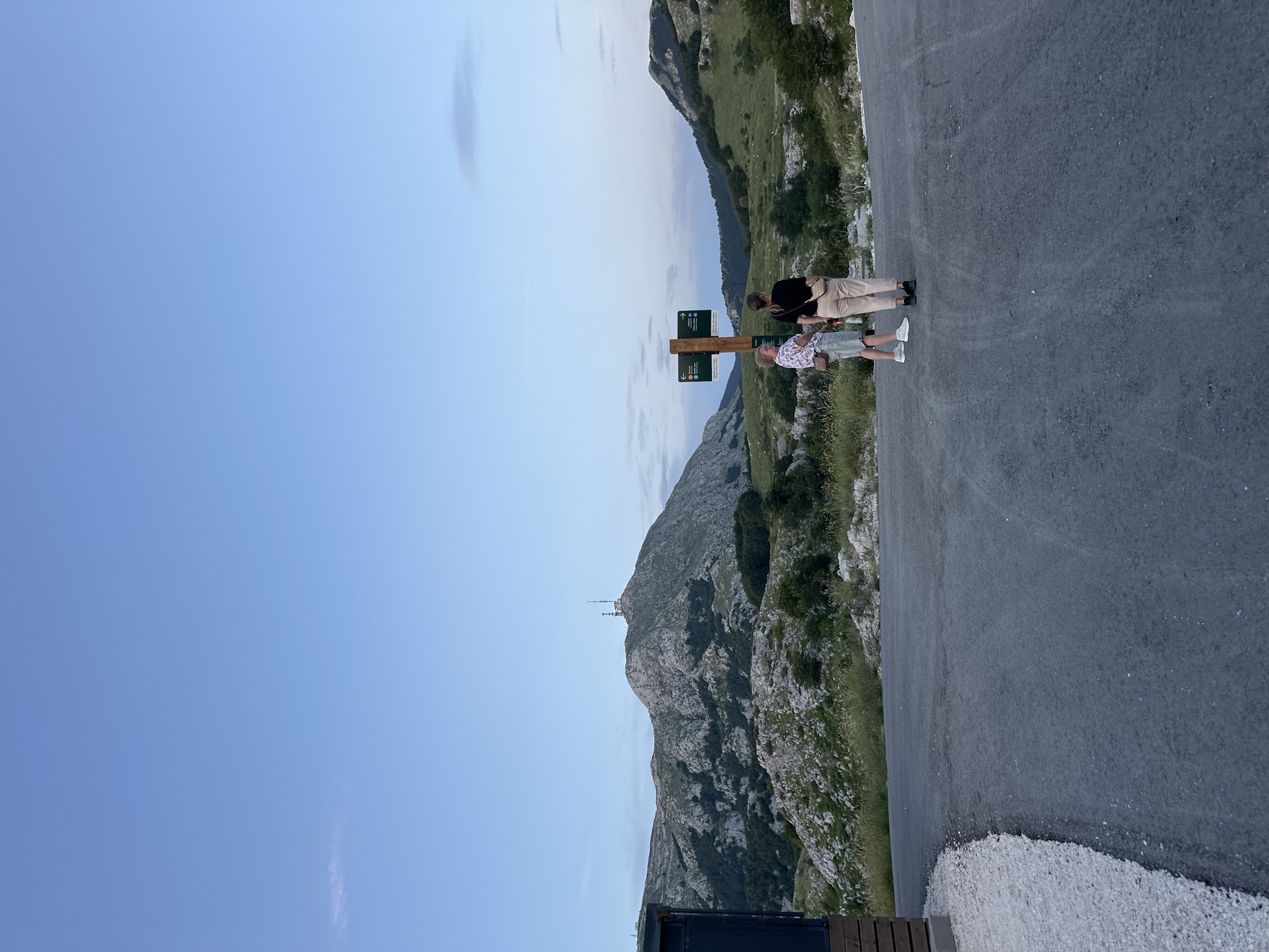 Two individuals stand near a wooden signpost with directional arrows against a backdrop of a mountainous landscape at dusk. The signpost indicates directions to "Jezerski Vrh" and "Mausoleum." The terrain is rocky with sparse vegetation and trees. A paved road leads away from the viewer towards the signpost. To the left, a dark building structure with wooden siding is partially visible. The sky is clear blue with a faint gradient indicating twilight. The overall scene suggests a viewpoint in a mountainous region, likely a national park or a scenic overlook.