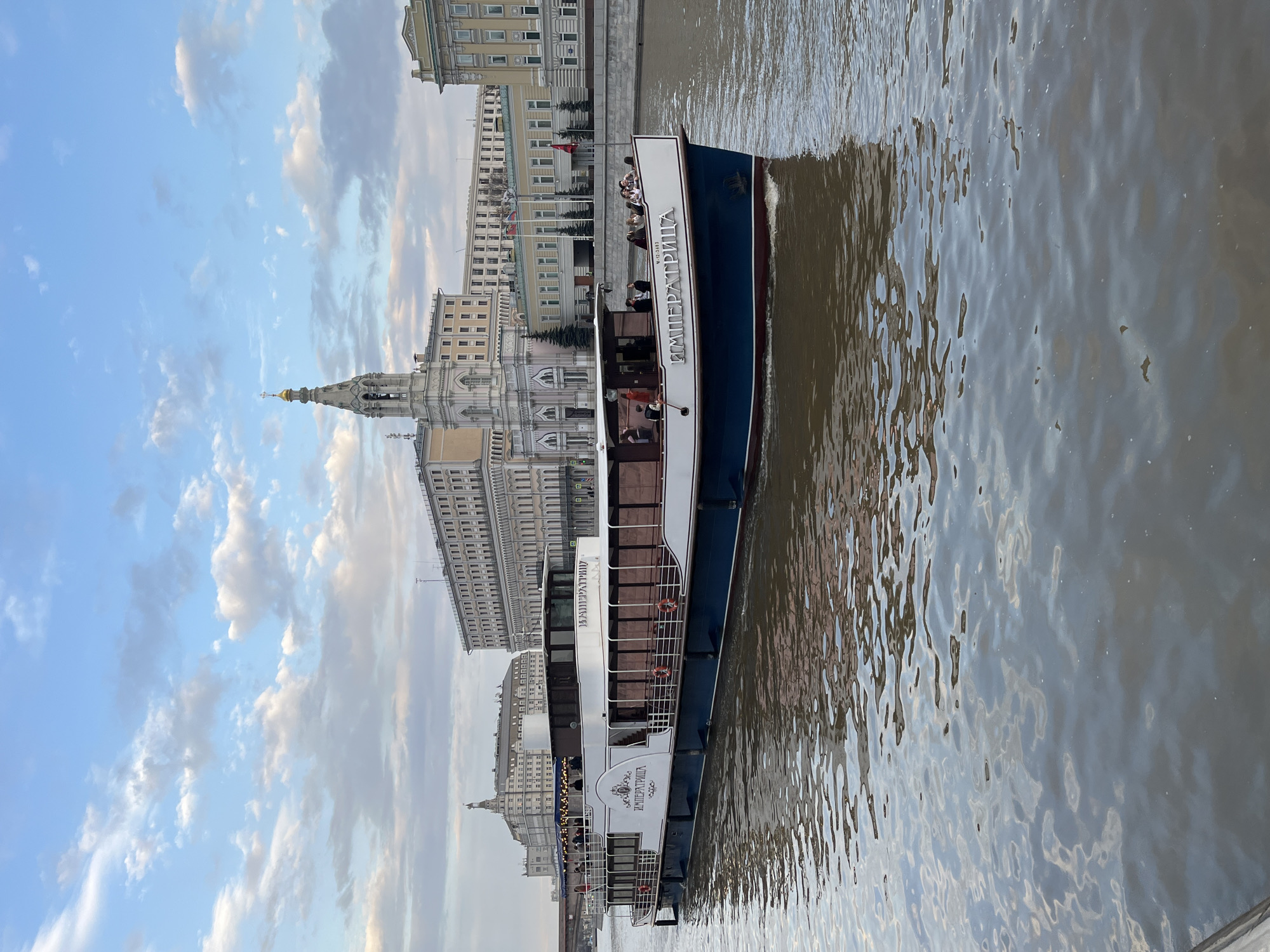 The image depicts a riverboat, named "Императрица", traveling on a river in Moscow, Russia. Passengers are visible on the boat's upper deck. The river reflects the surrounding buildings and sky. In the background, there are buildings typical of Moscow's architecture, including one with a prominent spire. The sky is filled with clouds. The river water is murky, reflecting the light.