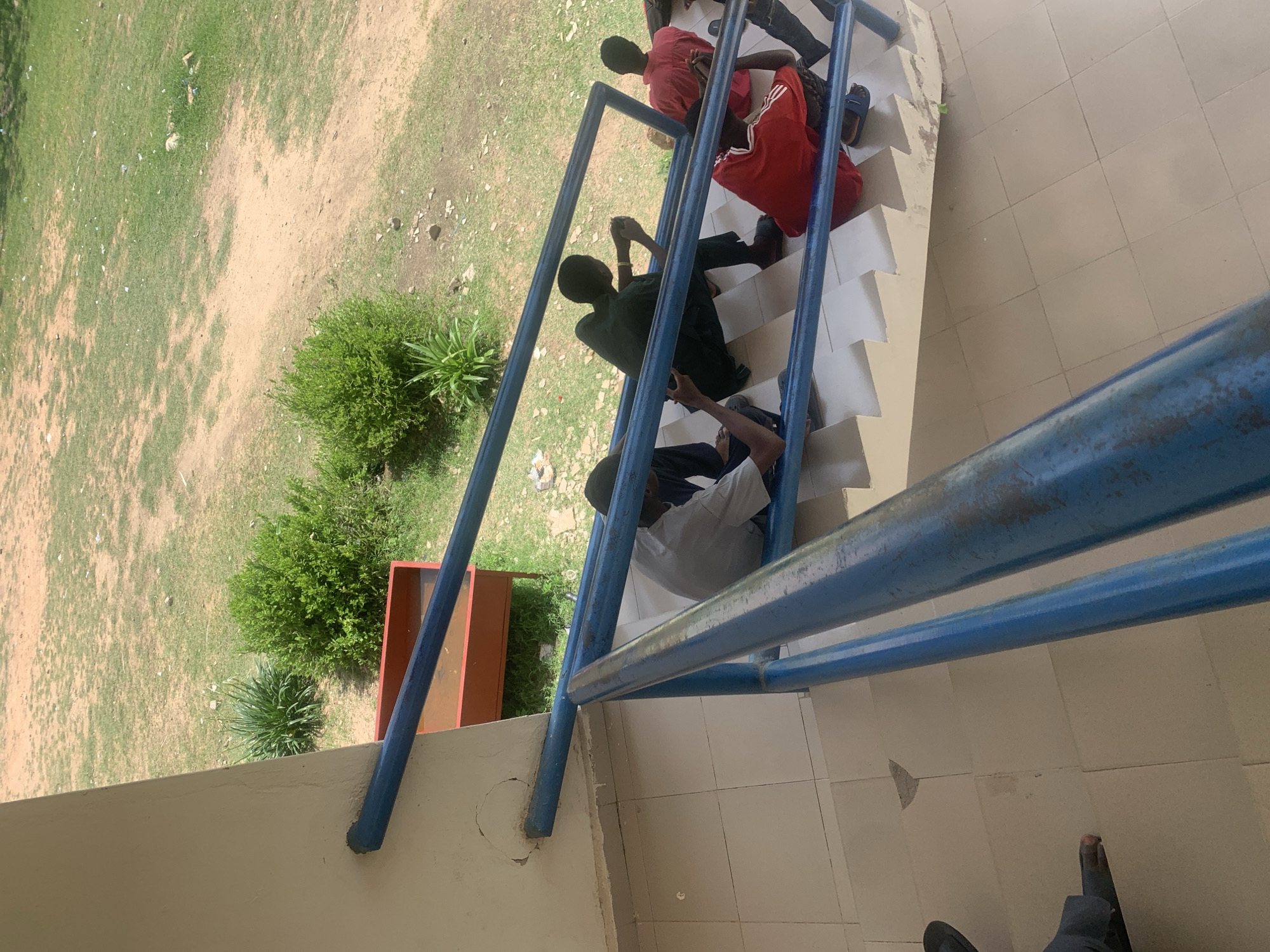 The image shows a group of young men sitting on a concrete staircase with blue railings. The staircase appears to be outdoors, with a grassy and sandy area visible in the background, possibly a campus or a public space. The young men are casually dressed, with some appearing to be using mobile phones. The overall scene suggests a relaxed atmosphere. Based on its appearance, this staircase could be located within the Federal University of Technology, Jalingo, in Jalingo, Nigeria. The architecture of the building and the surrounding vegetation are consistent with the typical environment of a university campus in that region.