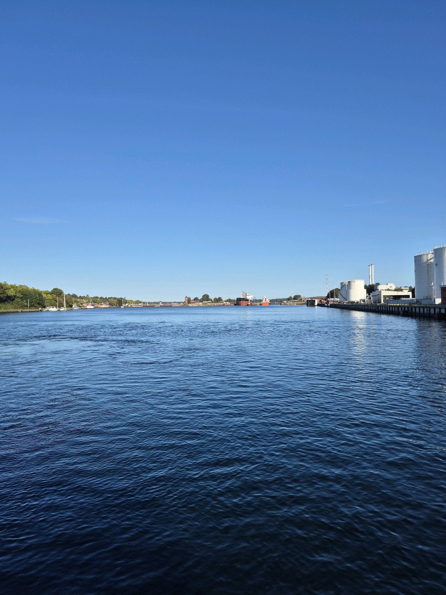 The image depicts a wide, calm body of water, likely a harbor or river, under a clear, bright blue sky. On the right side of the frame, a waterfront industrial area is visible, featuring several large, white cylindrical storage tanks, some with a number "08" visible on them, and various industrial structures including pipes and buildings. A long pier or dock extends into the water. Further in the distance, several large ships, some red and some dark, are anchored or moving in the water. The left side of the water shows a lush green tree-lined bank with some buildings and sailboats moored near the shore. The overall impression is a bustling port or industrial waterfront area. This scene is likely located in the city of Kiel, Germany, given the presence of industrial port facilities and maritime activity common to the city.