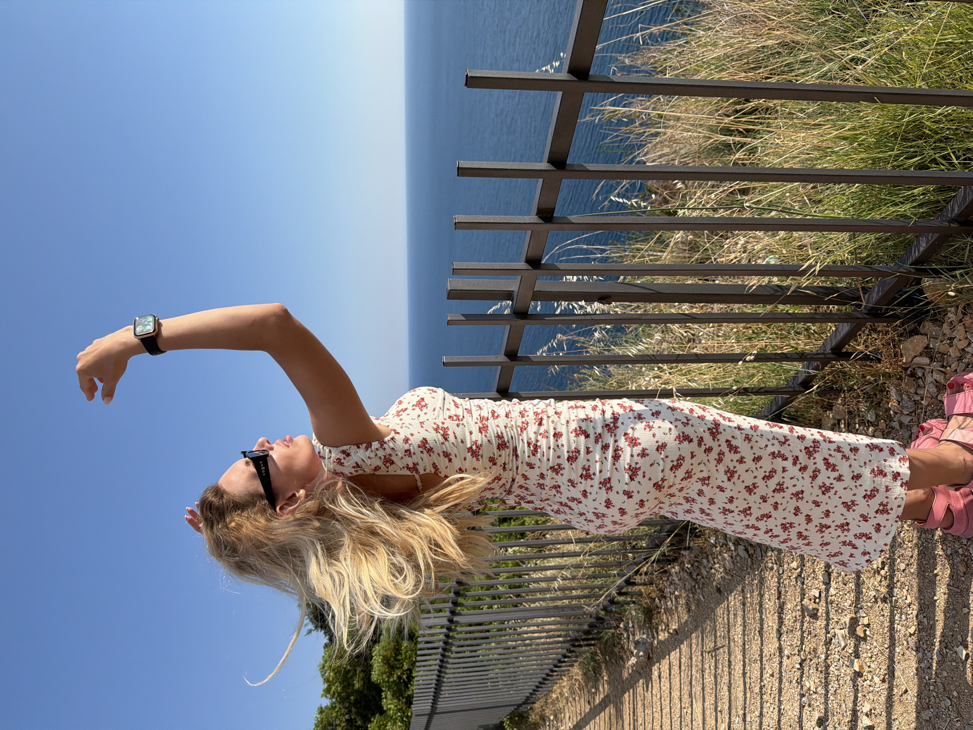 A blonde woman stands on a dirt path overlooking the Adriatic Sea in Prijevor, Montenegro. She is wearing a white floral print dress with red flowers, pink sandals, and black sunglasses. Her hair is blowing in the wind as she reaches her hand up towards the clear blue sky. A metal fence runs along the edge of the path, separating it from the grassy cliffside and the sea below. The sun casts shadows on the path, indicating it is late afternoon.