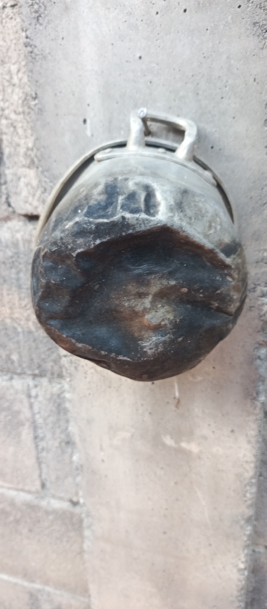 The image shows a close-up, slightly angled view of a dented, dark metal pot with a metal handle. The pot is attached to a textured, gray surface, likely a wall. The surface has visible mortar lines, suggesting it is made of brick or stone. The lighting is somewhat diffused, creating soft shadows and highlighting the distressed texture of the pot. There is no indication of the specific location within Jatisari, Indonesia, or any people or actions.