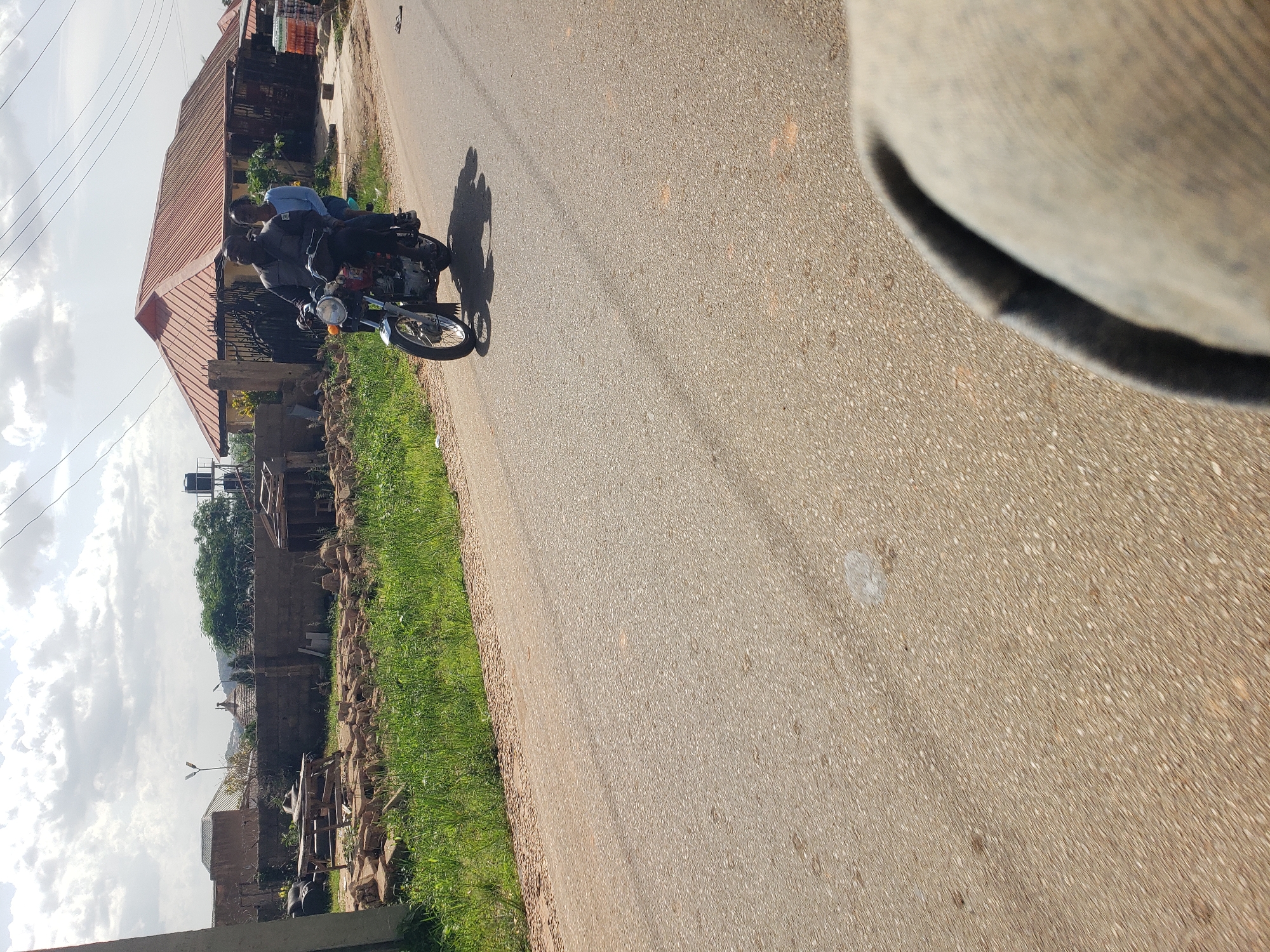 The image captures a street scene in Gura Top, Nigeria. A man and a woman are riding on a motorcycle. The man is driving, wearing a dark jacket, and the woman is seated behind him, wearing a light blue shirt. The motorcycle appears to be a red and chrome model. They are traveling on a paved road with grass and a low wall bordering one side. In the background, there are buildings with red-tiled roofs, some of which appear to be under construction or unfinished. Various materials, including bricks and wooden structures, are visible on the side of the road, suggesting ongoing construction or a storage area. The sky is partly cloudy, with sunlight casting shadows on the road. The perspective of the photograph suggests it was taken from another moving vehicle, with a close-up view of a person's knee in the foreground.