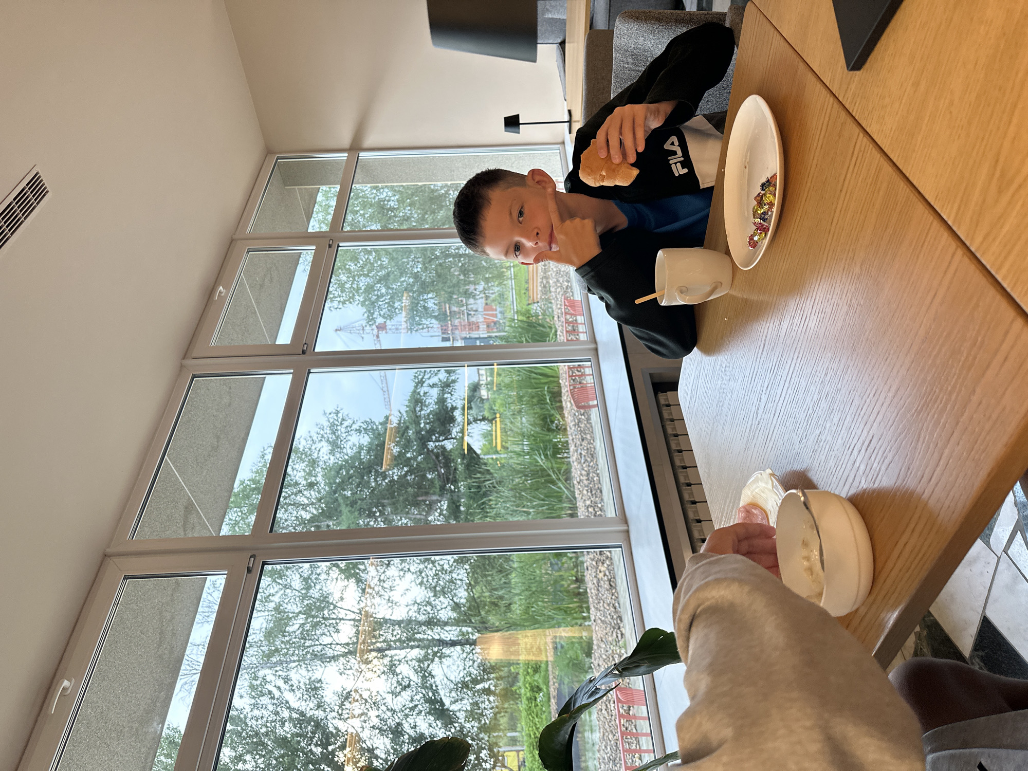 The image shows a young boy sitting at a wooden table in a brightly lit room with large windows. He is holding a piece of food, possibly bread, in his right hand and is gesturing with his left hand towards his cheek. He is wearing a dark jacket with white and black accents and a blue shirt underneath. In front of him on the table is a white mug with a wooden stirrer and an empty white plate with several colorful candies.
To the left of the boy, another person's arm and hand are visible, reaching towards a small white bowl containing what appears to be food, possibly oatmeal or porridge, and a small plate with slices of meat. The background through the windows shows trees, greenery, water, and construction cranes, suggesting an outdoor environment, possibly a park or an area undergoing development. The room appears to be a dining area or cafe. The flooring consists of a checkered pattern of black and white tiles. The lighting in the room is soft and ambient, coming from the large windows and possibly overhead fixtures not visible in the frame. The overall scene conveys a casual mealtime setting. The presence of construction cranes in the background suggests the location might be in an area of ongoing construction or development. This specific detail combined with the given location suggests the image might have been taken in Bolshoye Savino, Russia, a district known for its airport and surrounding development.
