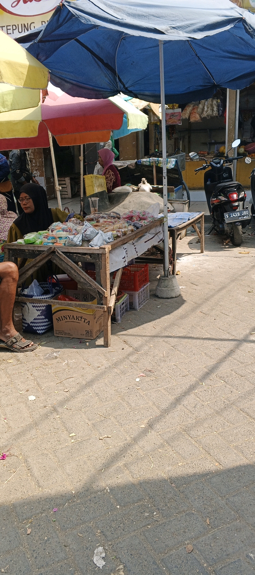 The image depicts a street scene in Waruwetan, Indonesia, likely at a market. Underneath a large blue awning, a woman wearing a black hijab and glasses sits behind a wooden cart laden with various packaged food items. To her right, another woman in a pink hijab stands. Several colorful umbrellas are visible, suggesting a covered market area. A black motorcycle is parked to the right of the cart, its license plate reading "S 2419 JBU". In the foreground, a cardboard box labeled "MINYAKITA" is placed on the ground. The ground is paved with interlocking bricks, and there are scattered leaves and debris. Shadows cast by the awnings and potentially other structures stretch across the paved surface, indicating it is a sunny day.