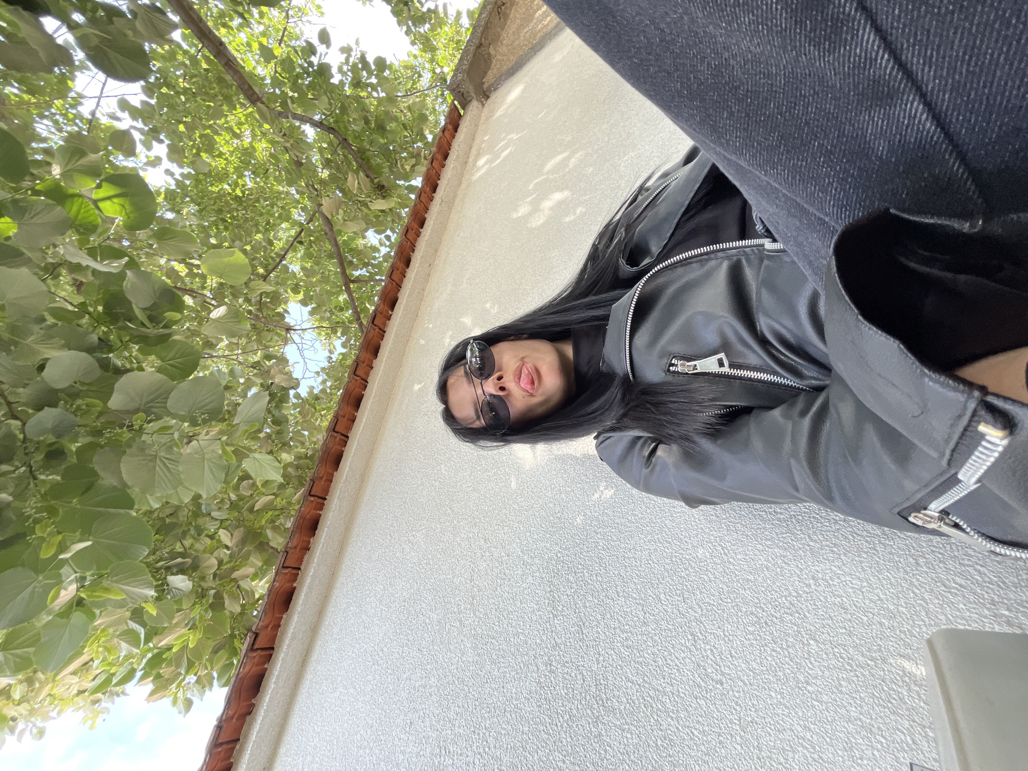 A woman is the primary subject, wearing a black leather jacket and dark jeans, positioned in front of a white textured wall. A tree with green leaves is visible in the background on the left. The scene appears to be outdoors, with glimpses of a red-tiled roof above the white wall. Given the stated location, the scene is in Saedinenie, Bulgaria.