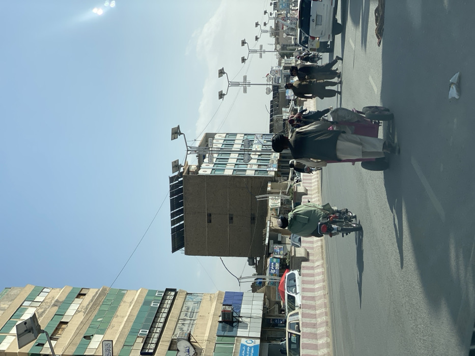 This image depicts an urban street scene in Kandahar, Afghanistan. The view is from a vehicle, looking down a multi-lane asphalt road under a clear, bright blue sky with a visible sun glare in the upper right.
In the foreground, the asphalt surface of the road is visible, with a small piece of white debris near the center-right. The road is divided by concrete barriers featuring alternating white and red/orange stripes.
Midground elements include active traffic. On the left lane, an individual wearing a light green poncho or cloak rides a motorcycle away from the camera. In the adjacent right lane, another individual, dressed in traditional attire including a dark turban, light vest, and light-colored shalwar kameez, pushes a two-wheeled pushcart with a red frame and grey container, also moving away. Further in the right lane, a white SUV is part of the traffic flow. Several pedestrians, dressed in traditional Afghan clothing, are visible walking or crossing the street on the far side of the road.
The background features multiple buildings and street infrastructure. On the left, a tall, multi-story building with a weathered facade is prominent, characterized by alternating bands of beige and teal/green paneling. Multiple windows are visible, along with signs featuring Arabic script on its lower floors. A street light fixture is mounted on this building. To the center-right, a shorter, blocky brown building, approximately four to five stories tall, has an array of solar panels mounted on its flat roof. Next to it on the right stands another multi-story building with blue-tinted windows or panels. The street is lined with numerous tall, metallic street light poles, each featuring a distinctive dual-lamp fixture and topped with a small solar panel, indicating a solar power source. These poles extend into the distance, delineating the street. A single dark cable spans horizontally across the upper-middle section of the frame. Long shadows are cast on the road, consistent with bright daylight.
