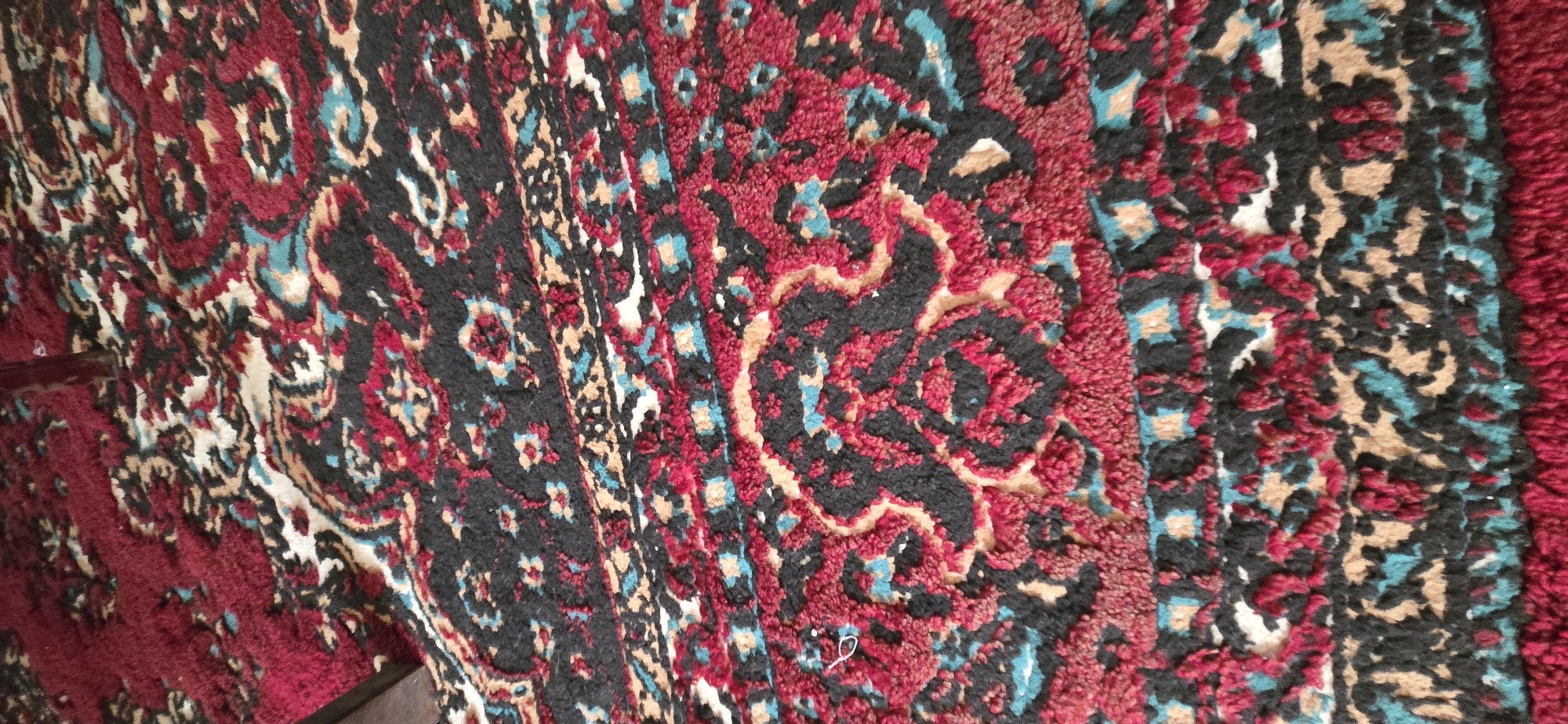 The image shows a close-up view of a patterned rug with a dark red background. The pattern is intricate and features swirling motifs in black, light brown, and a teal-blue color. There are also small white accents within the pattern. The rug appears to be made of a textured material, possibly with a high pile, as suggested by the fuzzy appearance of the fibers. The lighting in the image is somewhat dim, and the angle of the shot is from above, looking down onto the surface of the rug. The bottom left corner of the image shows a very small piece of a dark brown object, possibly furniture, encroaching into the frame. There are no people or animals visible in the image. The rug is lying flat on a surface. This image is not sufficient to determine any specific location in Tel, Azerbaijan.