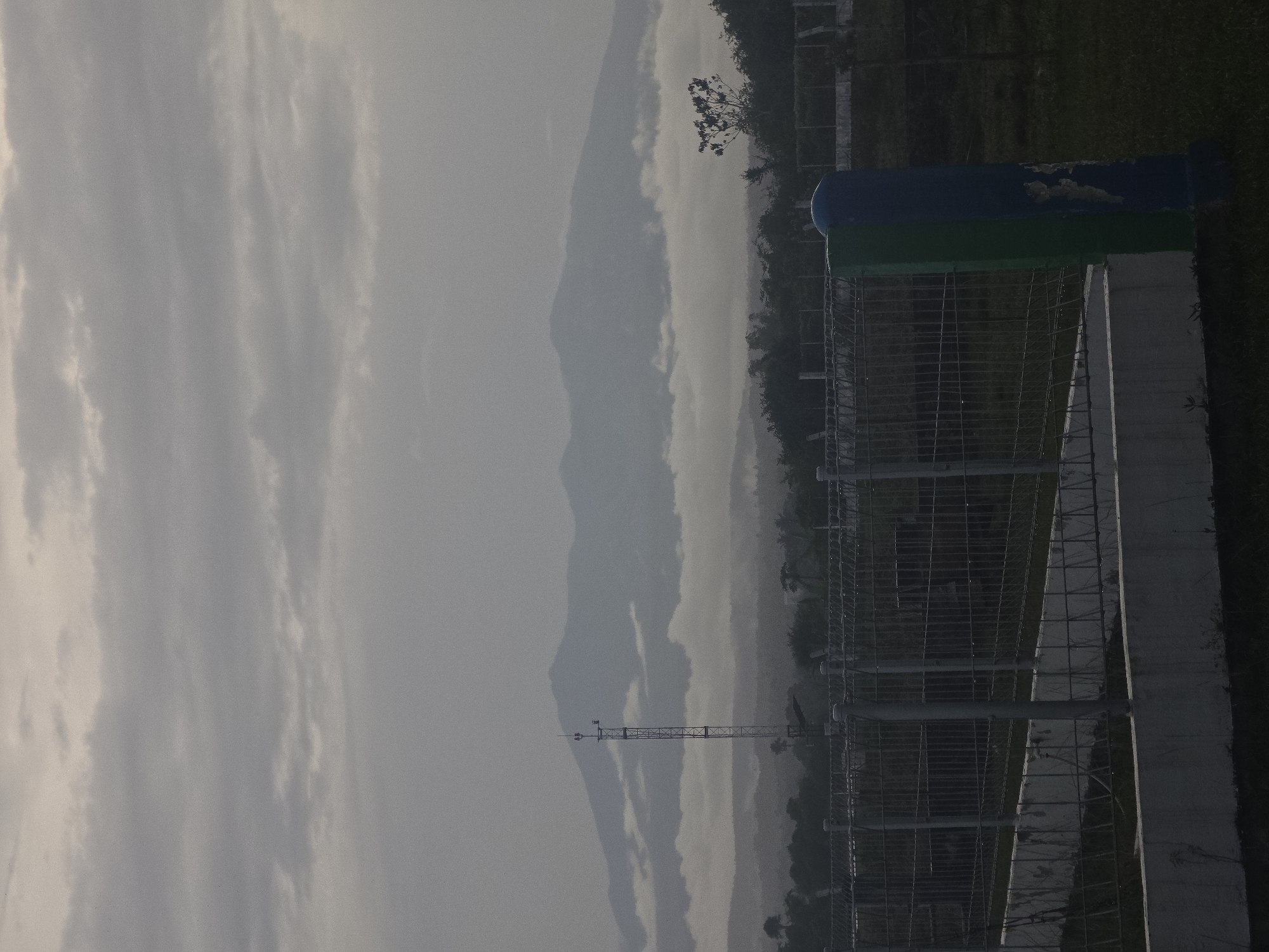 The image depicts a landscape in Pintomakmur, Indonesia, under an overcast sky. Distant, hazy mountains are visible. In the foreground, there's a section of a metal fence structure. Adjacent to the fence is a concrete structure, next to which is a blue and green cylindrical object. A thin communication tower stands to the left of the fence. The environment is lush with vegetation, including trees and grass.