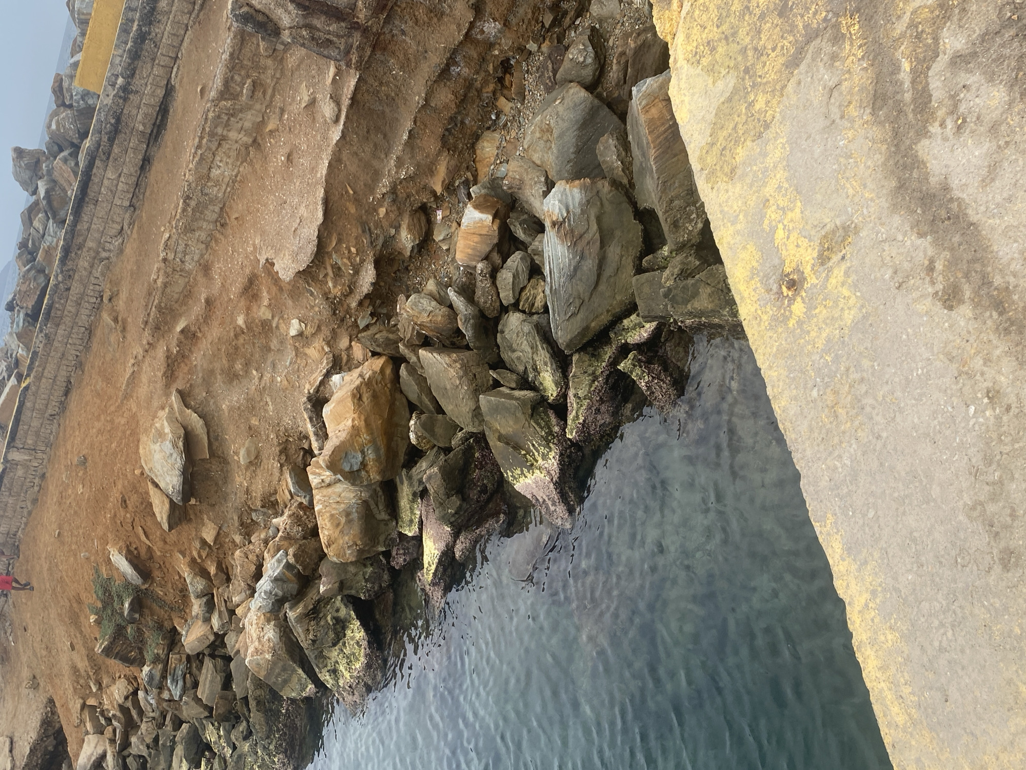 The image shows a rocky shoreline in Porlamar, Venezuela. A concrete structure is visible on the right and top-left of the image, suggesting the edge of a man-made construction, possibly a pier or seawall. The shoreline consists of dirt, sand, and various sizes of rocks and boulders, some extending into the water. The water is calm and relatively clear, with some visible details beneath the surface. A small bush is visible on the shoreline.