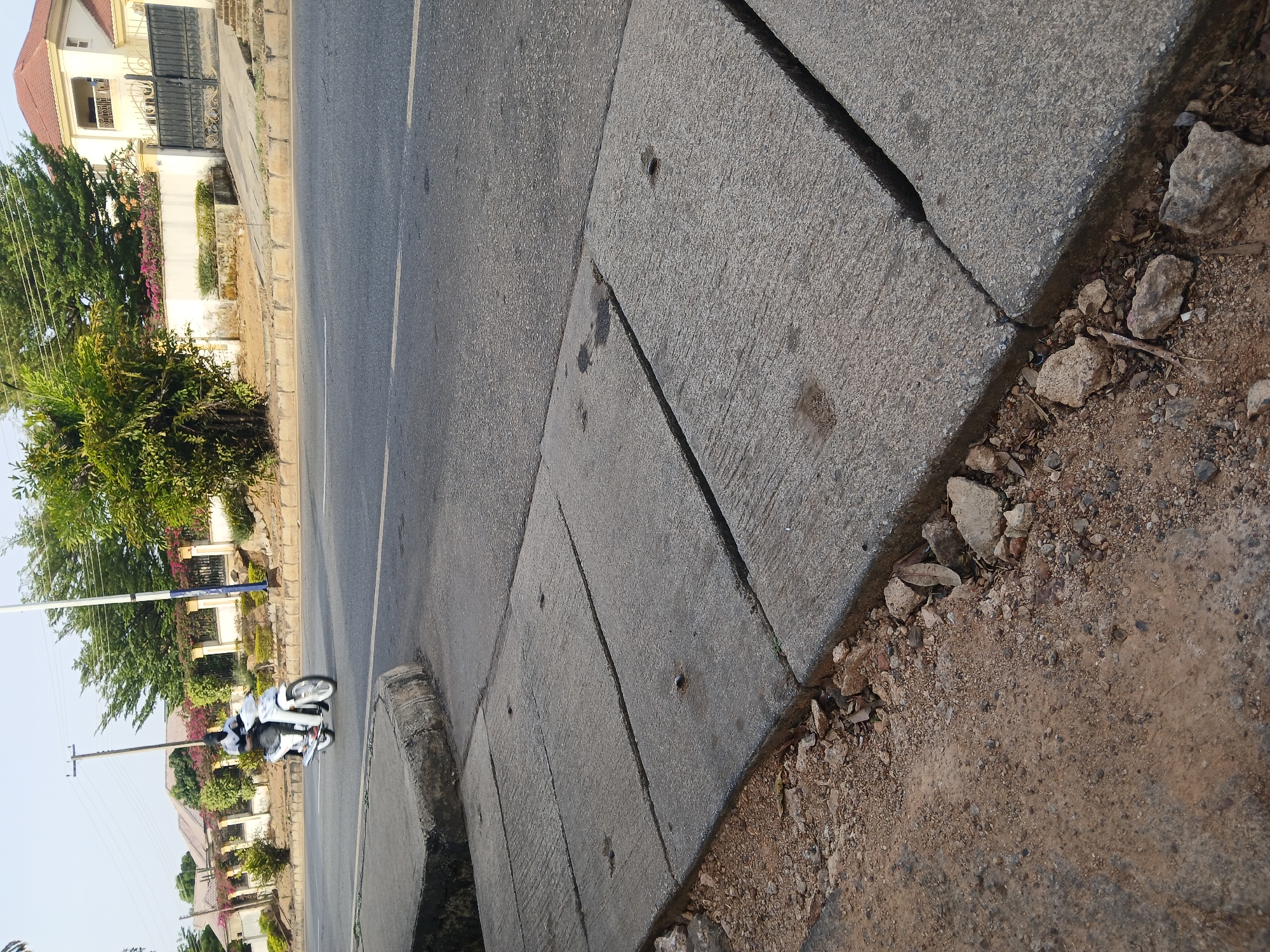 A street scene in Jos, Nigeria is depicted. The viewpoint is along a sidewalk made of concrete slabs, which show cracks and are bordered by loose dirt and gravel. A street runs parallel to the sidewalk, and a person on a white motorcycle is traveling on the road. Buildings and lush green vegetation are visible on the other side of the street. A utility pole is also visible.