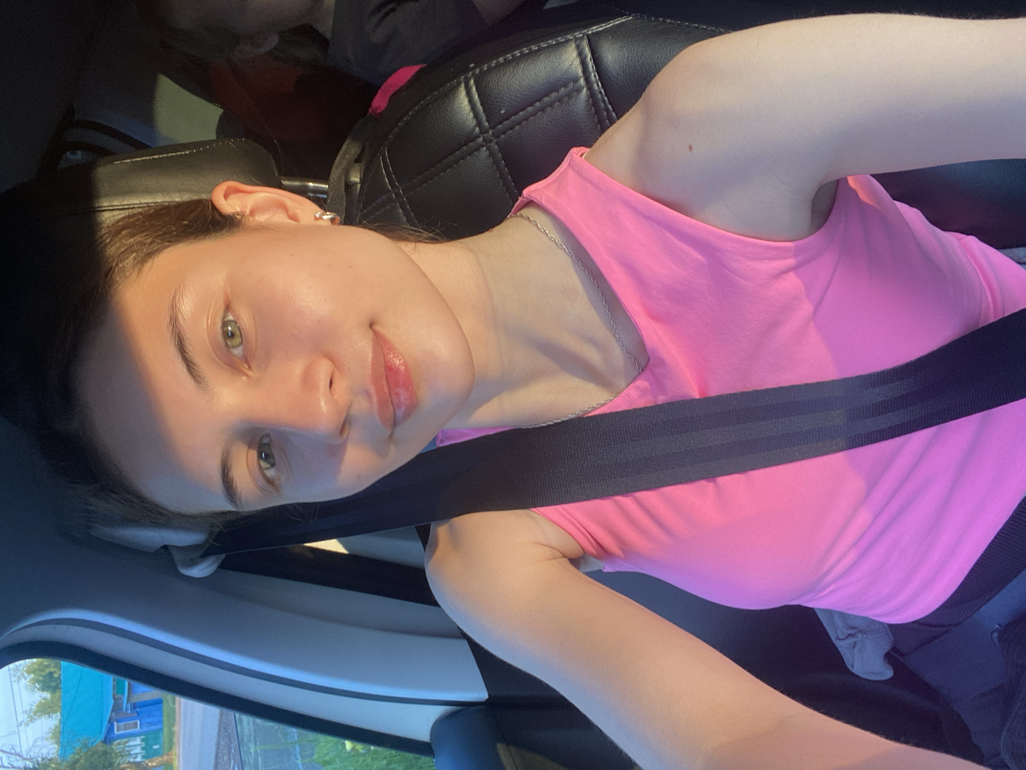 The image shows a young woman in the front passenger seat of a car, wearing a pink tank top and a seatbelt. She has light brown hair and is looking towards the camera with a slight smile. Sunlight is casting shadows on her face and upper body. The interior of the car is visible, including a black leather seat and part of another person in the back seat. Outside the car window, a rural scene is visible with a blue house and green trees. This image was captured in Votkinsk, Russia, specifically on the street of Komsomolskaya, near house number 14, as indicated by the background details.