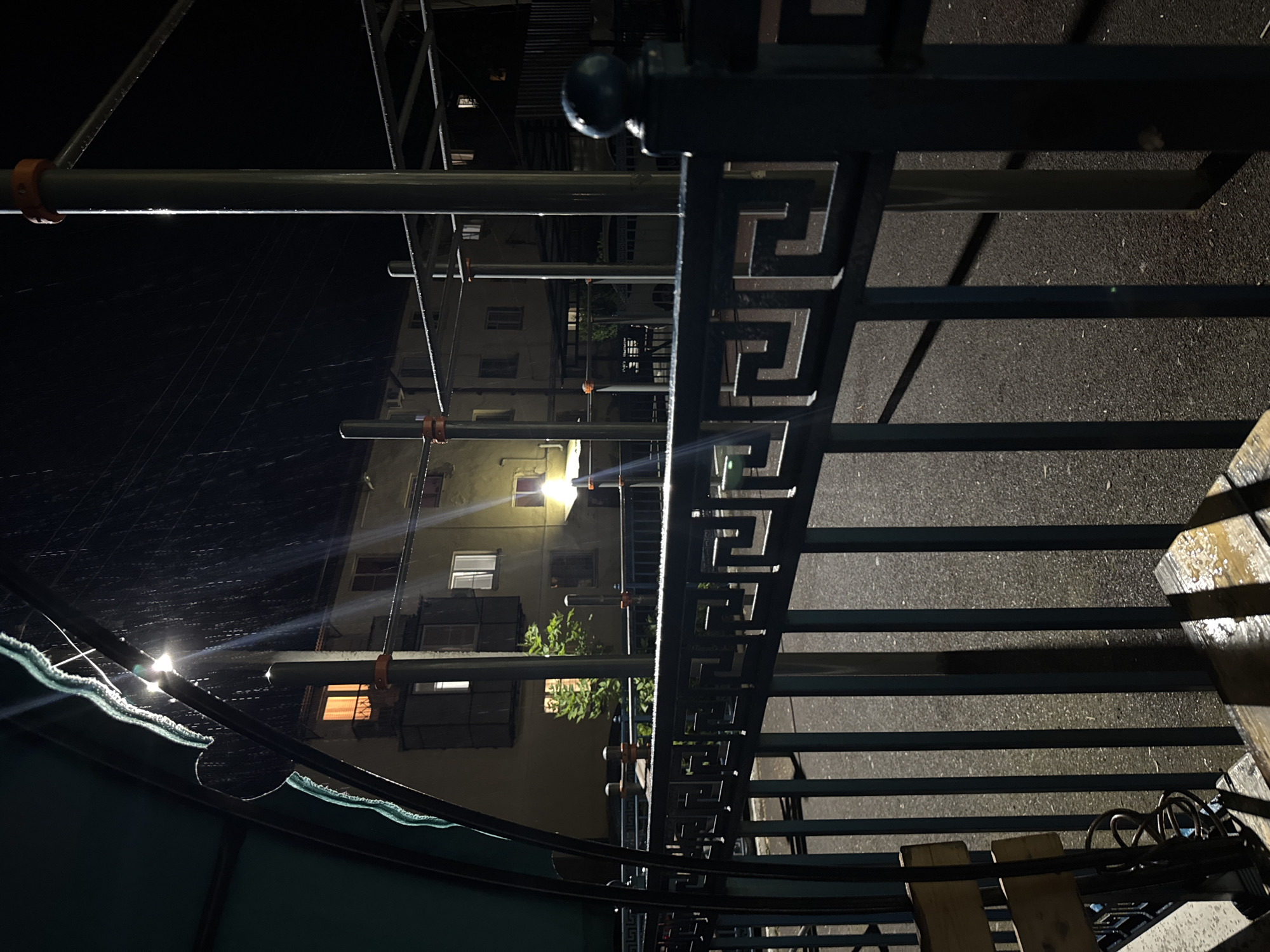 A nighttime, outdoor scene at an urban sports ground with visible rain falling. In the foreground, a dark metal fence with a Greek key pattern separates the viewer from the rest of the scene. A portion of a wooden bench is visible on the left, and what appears to be a bicycle lock is attached to a metal pole. Behind the fence, a sports ground with parallel bars and upright poles is visible. The ground surface is asphalt and wet from the rain. In the midground and background, a multi-story building with lit windows is present. The overall lighting is dim, with artificial lights illuminating the sports equipment and the building. The rain creates visible streaks of light as it falls through the artificial illumination.