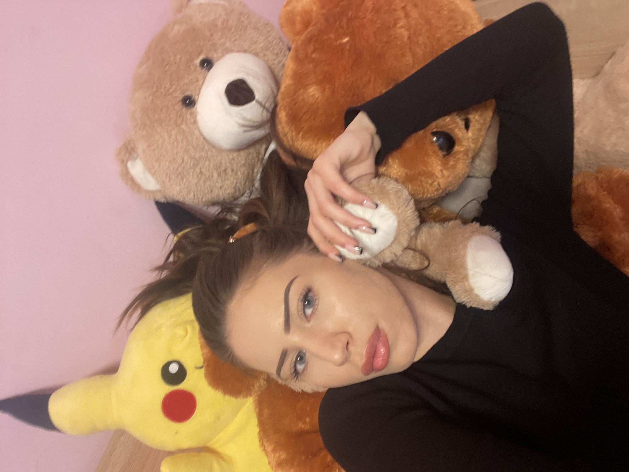 A woman lies among stuffed animals, including a Pikachu and several teddy bears. The background is a plain, light pink wall. She wears a black long-sleeved shirt. The location is Somovit, Bulgaria.