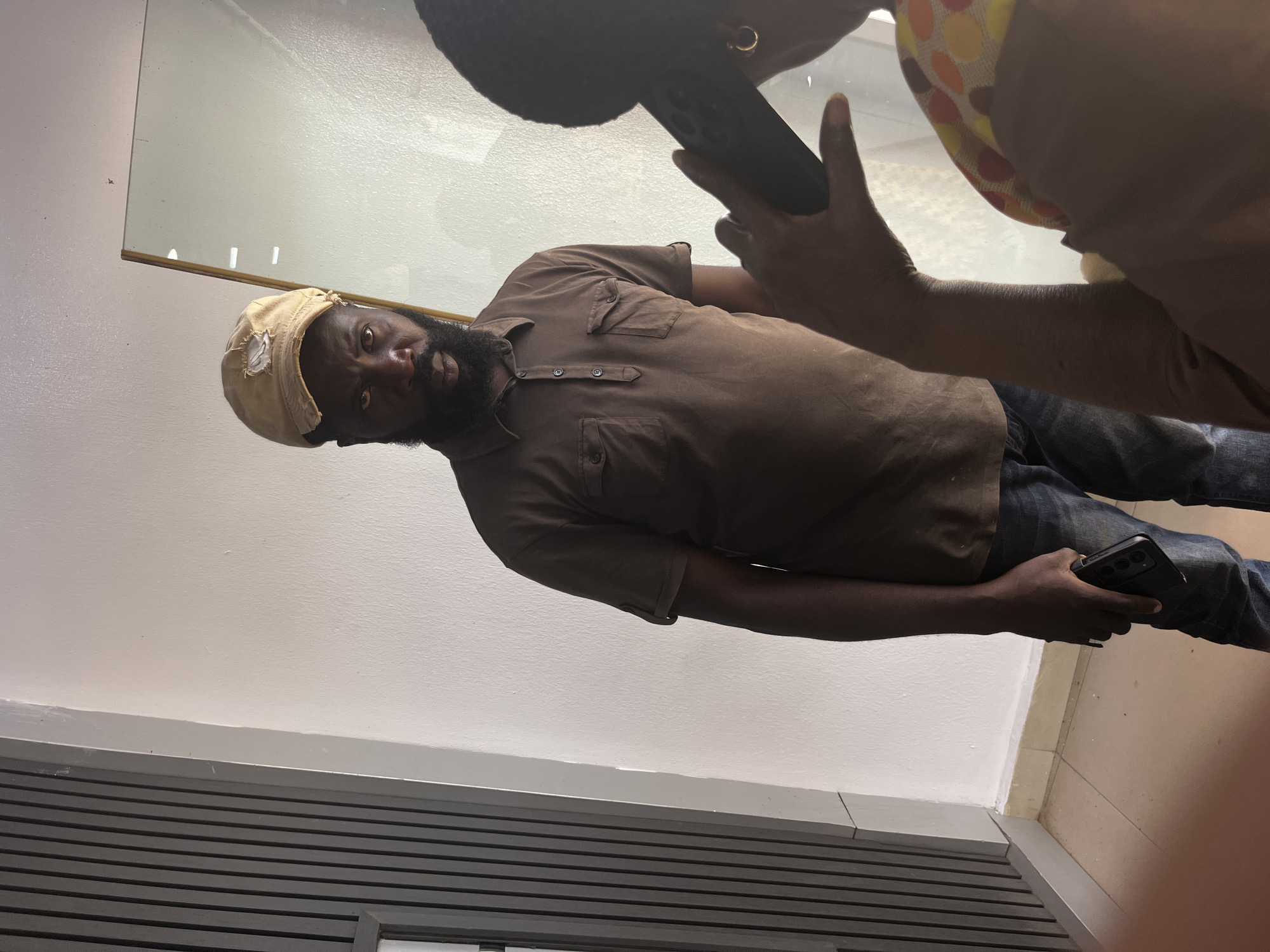 A man stands in a room with a light-colored wall and a ribbed, dark gray vertical panel on the left. He is wearing a tan baseball cap with a torn front, a brown collared polo shirt with two chest pockets, and dark blue jeans. He has a beard and is holding a dark-colored smartphone in his right hand. In the foreground, partially obscuring the man, is a person from behind, their left arm extended as they hold a dark-colored object to their ear, possibly a phone. This person is wearing a colorful, patterned garment and a gold hoop earring. The scene appears to be indoors, possibly a residential or commercial space, given the interior wall finishes and the presence of a mirror reflecting the scene. The location in Gura Top, Nigeria, is not visually evident from the image itself.