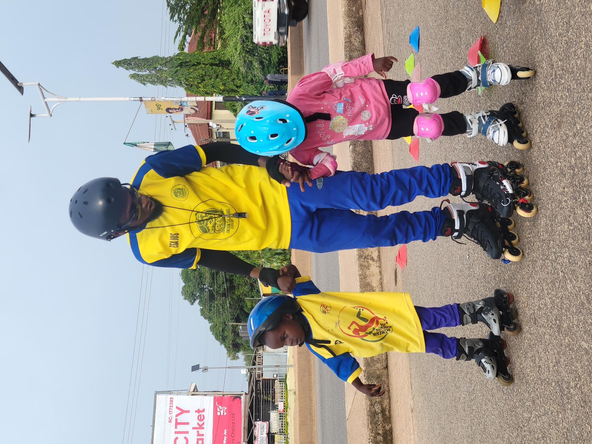In Jos, Nigeria, a man in a yellow and blue uniform, wearing a black helmet, is teaching two young children to rollerblade. He is holding the hands of both children, who are also wearing helmets and protective gear. The child on the left wears a blue helmet and a yellow shirt with blue sleeves, while the child on the right has a blue helmet and a pink long-sleeved shirt. The scene takes place on an asphalt surface, likely a street or an open area, with a roadside in the background. Traffic cones in various colors are scattered on the ground, suggesting a training or practice area. A billboard for "City Market" is visible in the background, along with trees, buildings, and a white Toyota truck driving on the road. The sky is overcast.