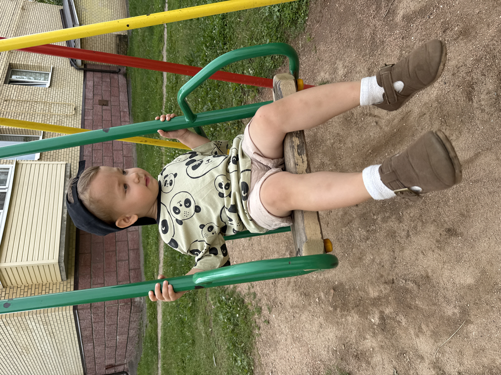 In Rusanovo, Russia, a young boy is seated on a wooden swing. He is wearing a light-colored t-shirt with a panda print, beige shorts, white socks, and brown shoes. A black baseball cap is on his head. The swing set has green, red, and yellow poles. The ground beneath the swing is sandy dirt, and there is green grass and a building in the background.
