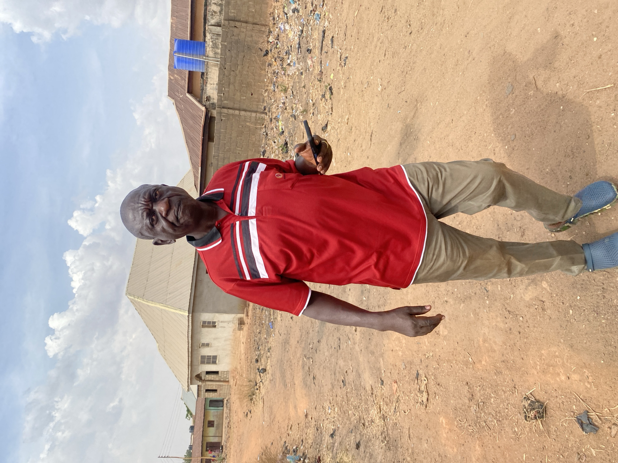 A male individual is depicted walking towards the viewer on a dirt road in a residential area of Rido, Nigeria, under a partially cloudy sky. He is an adult with dark skin, a bald head, and appears to be in his late middle age or early senior years. His facial expression is neutral to slightly stern, with his brow furrowed. He wears a red polo shirt with black and white horizontal stripes across the chest and shoulders, khaki-colored trousers, and blue perforated slip-on shoes. His right hand is down by his side, while his left hand holds a black smartphone horizontally.
The ground is unpaved, consisting of reddish-brown dirt with scattered dry vegetation and debris, including plastic bags and other refuse, particularly on the right side of the frame beyond a low concrete block wall.
In the background, on the left, a power pole with wires is visible, along with several single-story buildings in various states of construction or disrepair. One building to the left has a light brown corrugated metal roof and what appears to be a light green painted facade. Another building further back has a white facade. Directly behind the subject, a partially constructed concrete building with unpainted walls and window openings features a light tan corrugated metal hip roof. To the right of this building, a low, unfinished concrete block wall extends horizontally. Beyond this wall, to the far right, another building with a dark brown corrugated metal roof and a prominent blue cylindrical water storage tank on its roof is visible. The overall scene suggests a developing urban or semi-urban environment.
