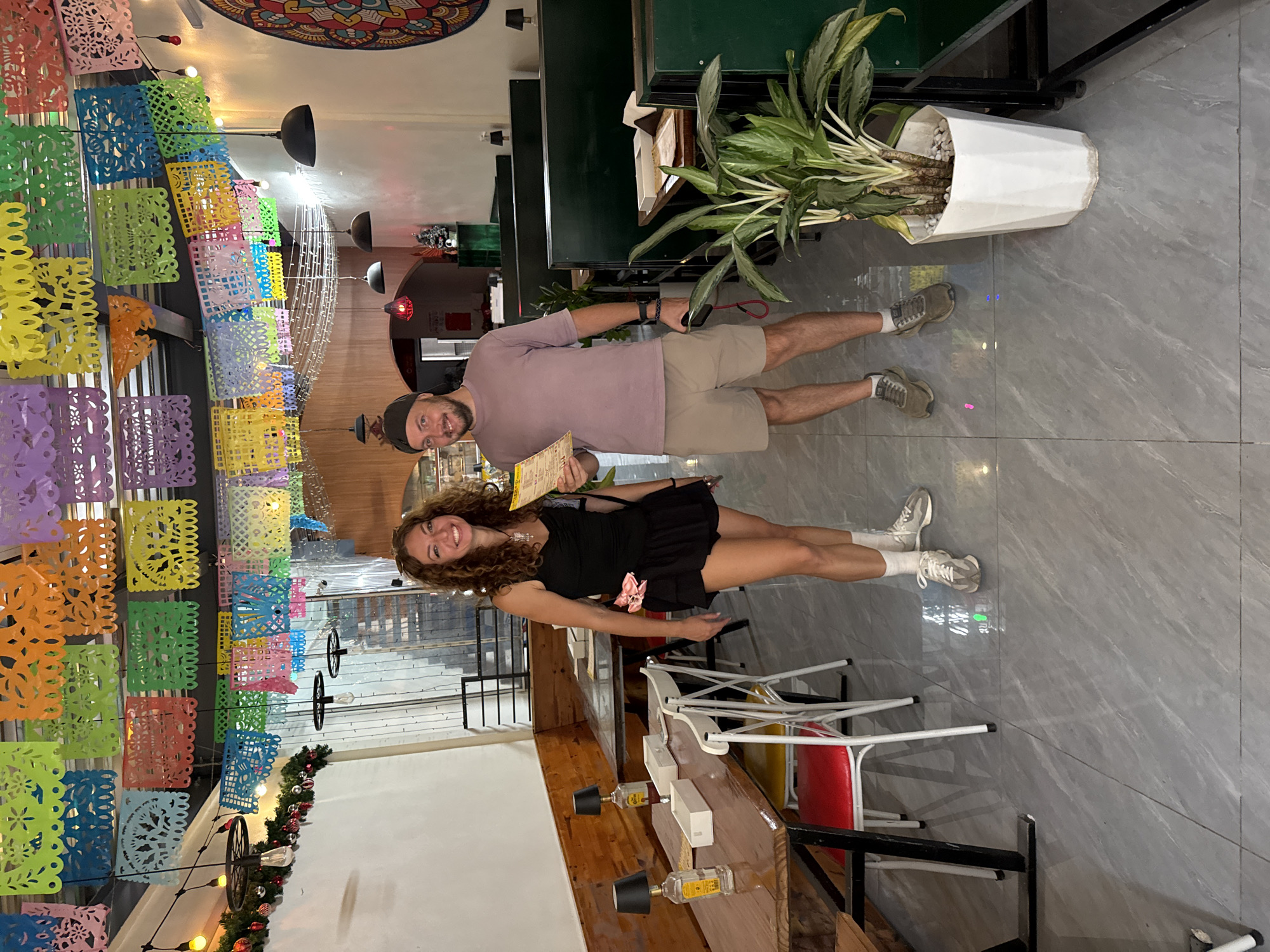 A medium shot captures a man and a woman standing in what appears to be a restaurant in Hoa Lư Ward, Vietnam, decorated with colorful papel picado banners and festive lights. The woman, on the left, is wearing a black sleeveless top and a black ruffled skirt with white socks and grey sneakers. She has curly brown hair and is smiling, holding a menu in her right hand. The man, on the right, is wearing a light purple t-shirt, tan shorts, white socks, and grey sneakers. He is also smiling and holding a menu in his left hand. They are positioned in the center of the frame, facing the camera.
The environment features wooden tables and green booths with lamps and potted plants. The floor is tiled with a grey marble pattern. In the background, more seating areas, lighting fixtures, and decorative elements like a mandala-style art piece are visible. The overall impression is a vibrant and welcoming atmosphere.