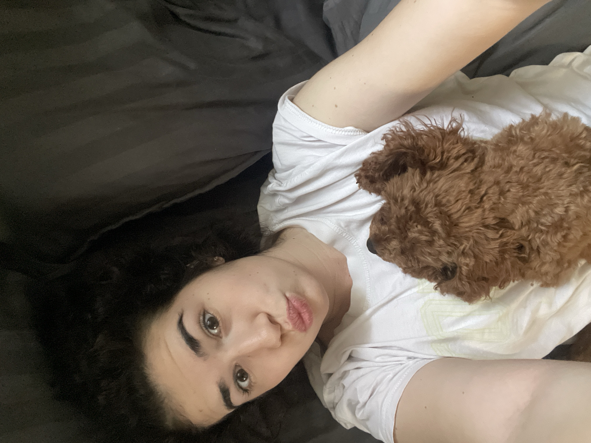 A woman lies on a dark gray bed, likely in a home or apartment, with a small, brown, curly-haired dog nestled on her chest. She is wearing a white t-shirt and has dark, curly hair. The location of the scene is not discernible from the image.