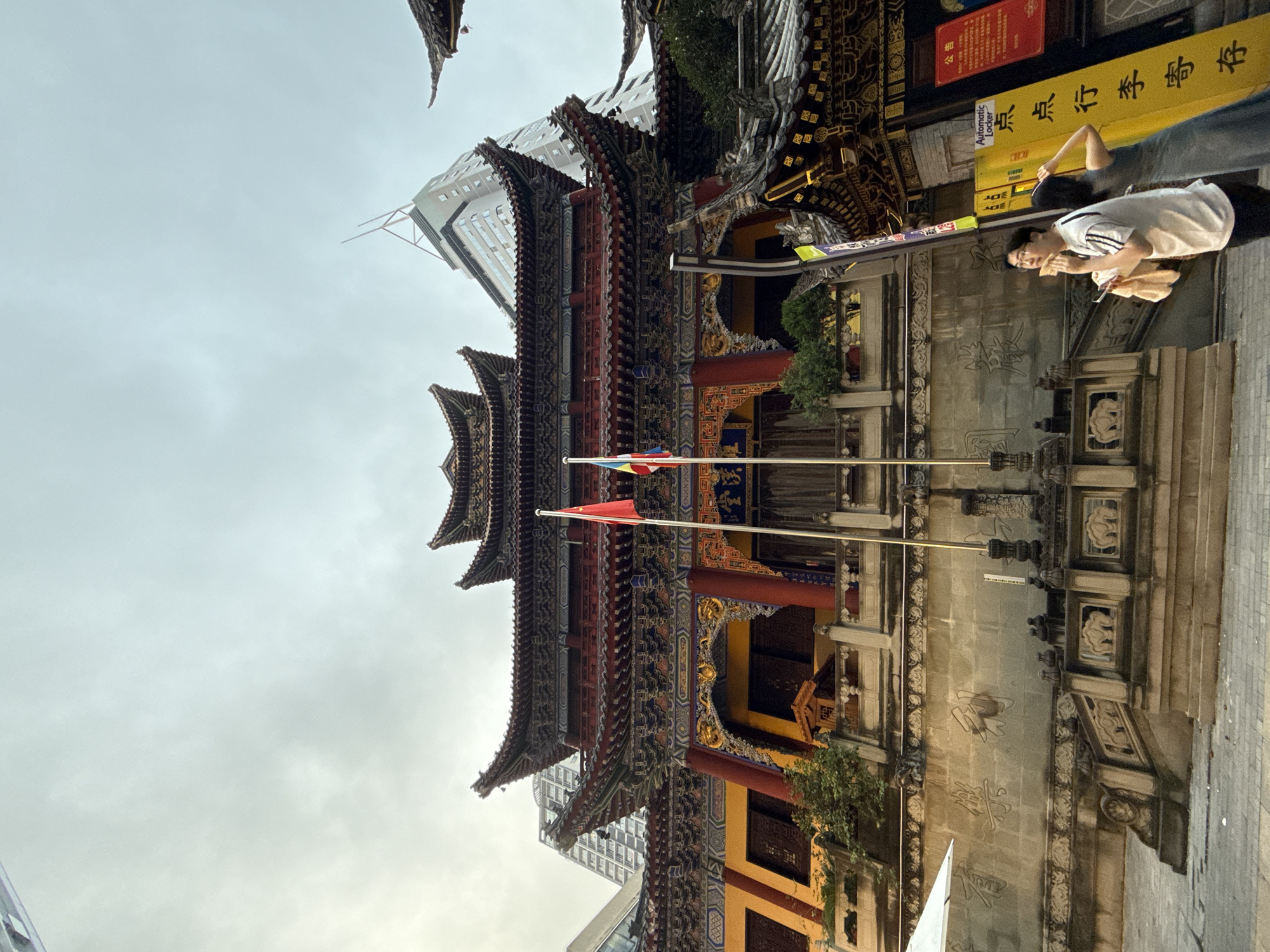 In Yuzhong District, China, a traditional Chinese temple with elaborate red and gold architecture stands prominently. The temple features tiered, curved roofs adorned with intricate carvings. Two flags, one red and one with multiple colors, fly from poles in front of the structure. A modern skyscraper looms in the background, contrasting with the ancient temple. In the foreground, two individuals are visible. One person, wearing a white t-shirt with black stripes and glasses, is holding a piece of food and a cloth, looking upwards. Another person, wearing a blue dress, stands behind them with their arm raised, appearing to adjust their hair. A yellow sign with Chinese characters and the words "Automatic Locker" is positioned to the right of the individuals. The ground is paved, and a stone wall with carved motifs and what appear to be elephant sculptures forms the base of the temple's elevated platform. The sky is overcast.