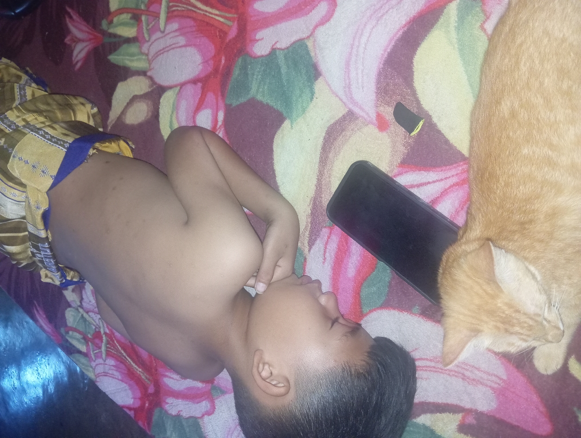 A young boy is lying down on a colorful blanket with a floral pattern. He is wearing a sarong with a yellow and brown plaid design and a blue waistband. His eyes are closed, and his hand is resting on his chin, suggesting he is sleeping or resting. Next to him, an orange cat is also lying down, seemingly asleep or relaxed. A dark-colored smartphone is placed on the blanket near the boy and the cat. The scene appears to be indoors, possibly a bedroom, given the blanket and the presence of personal items like a smartphone. The lighting suggests it could be daytime, though the exact time is not discernible. The overall mood is peaceful and intimate, capturing a moment of quiet rest shared by the boy and his pet. The floral pattern on the blanket is vibrant, with pink, white, and green elements. No text is visible in the image. The location context is Rejosari, Indonesia.