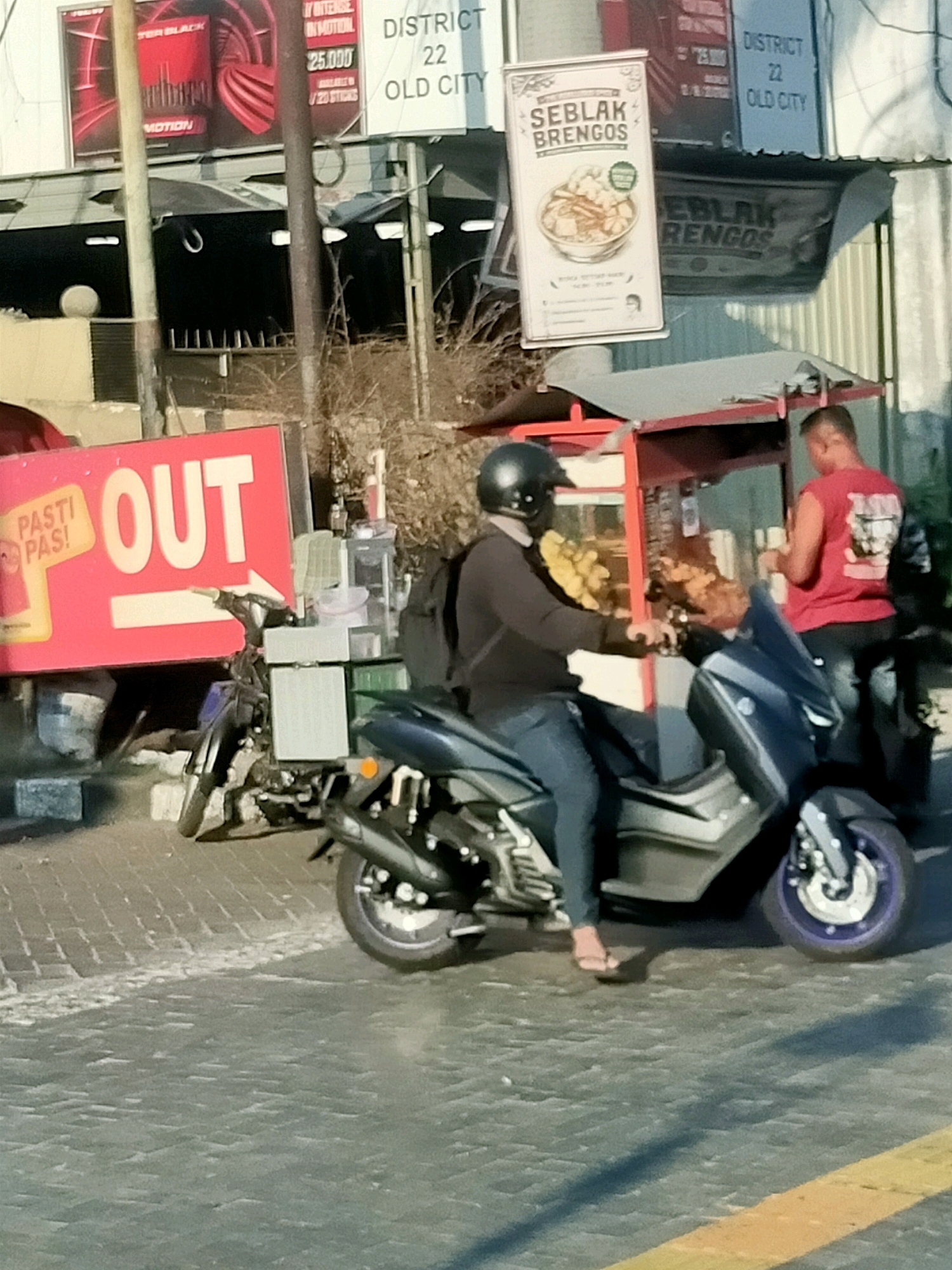 The image captures a street scene in Surabaya, Indonesia, likely in the Old City district, as indicated by signage. In the foreground, a person wearing a black helmet and dark clothing is seated on a dark-colored scooter, facing a food cart. The scooter has a blue windshield and the rider's legs are visible, wearing blue jeans and flip-flops.
The food cart is red and appears to be a mobile vendor. It has glass enclosures displaying food items, and a sign above it reads "SEBLAK BRENGOS," featuring an image of a bowl of food. Behind the cart, another individual wearing a red sleeveless shirt is visible, seemingly interacting with the vendor.
To the left of the food cart, a large pink sign with white text proclaims "PASTI PAS! OUT" with a white arrow pointing to the right. Further to the left, a black-and-white pole stands prominently. Various other signs are visible in the background, including one with "DISTRICT 22 OLD CITY" and another with text in Indonesian. The ground is paved with cobblestones. In the background, other vehicles and structures suggest a bustling urban environment.