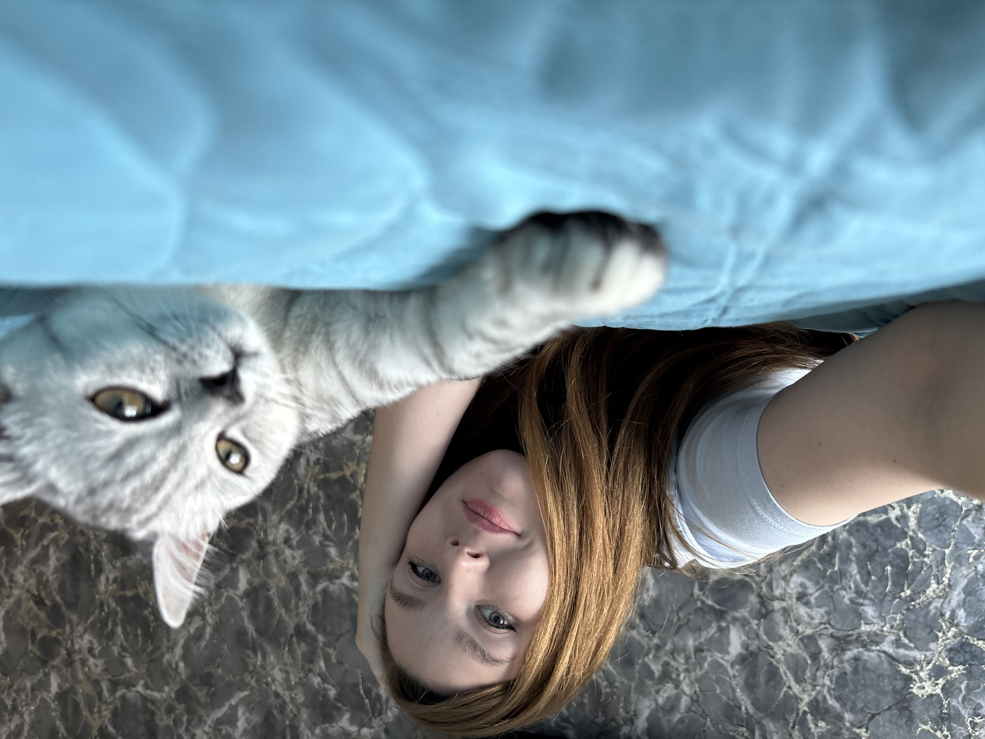 A young woman with long auburn hair is lying on a bed with a light blue blanket, taking a selfie. She is resting her head on her hand and smiling at the camera. Next to her, a silver tabby cat is also lying on the bed, with its head and paw reaching towards the camera. The background is a dark, marbled wall. The scene appears to be indoors, in a domestic setting. There is no information available to determine the specific location in Izhevsk, Russia.