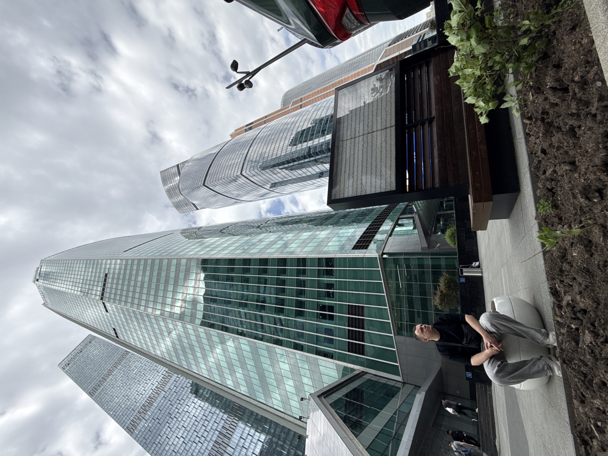 A low-angle shot captures a modern urban scene in the city of Unknown, Unknown, likely a financial district. Dominating the frame are several skyscrapers with reflective glass facades, towering against a cloudy sky. A person sits on a white, rounded seat near the bottom right of the image, dressed in a dark shirt and light gray pants. Adjacent to the person is a planter with greenery. A dark-colored vehicle is partially visible at the top of the frame. A modern bus stop is located near the planter. Other pedestrians are seen walking in the background. The image is slightly rotated.