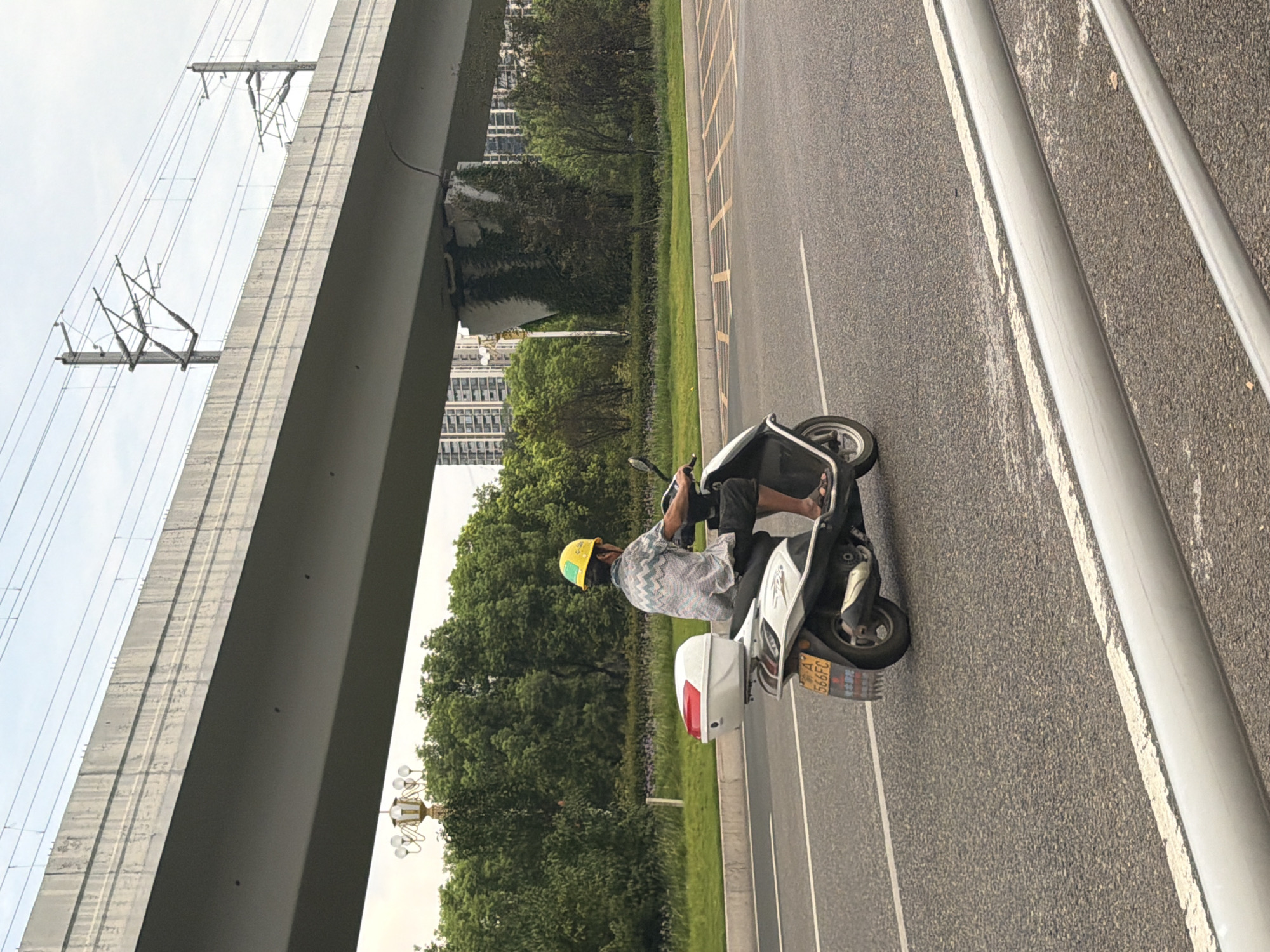 A person wearing a yellow hard hat rides a white and black scooter on an asphalt road. The scooter has a white storage box on the back. The rider is wearing a light-colored short-sleeved shirt with a pattern and dark shorts. In the background, there is a large concrete overpass with train tracks on top, surrounded by lush green trees and tall buildings. The sky is overcast. The license plate on the scooter is partially visible, showing "A 566FC". The location is identified as the city of Jiangjin, China.