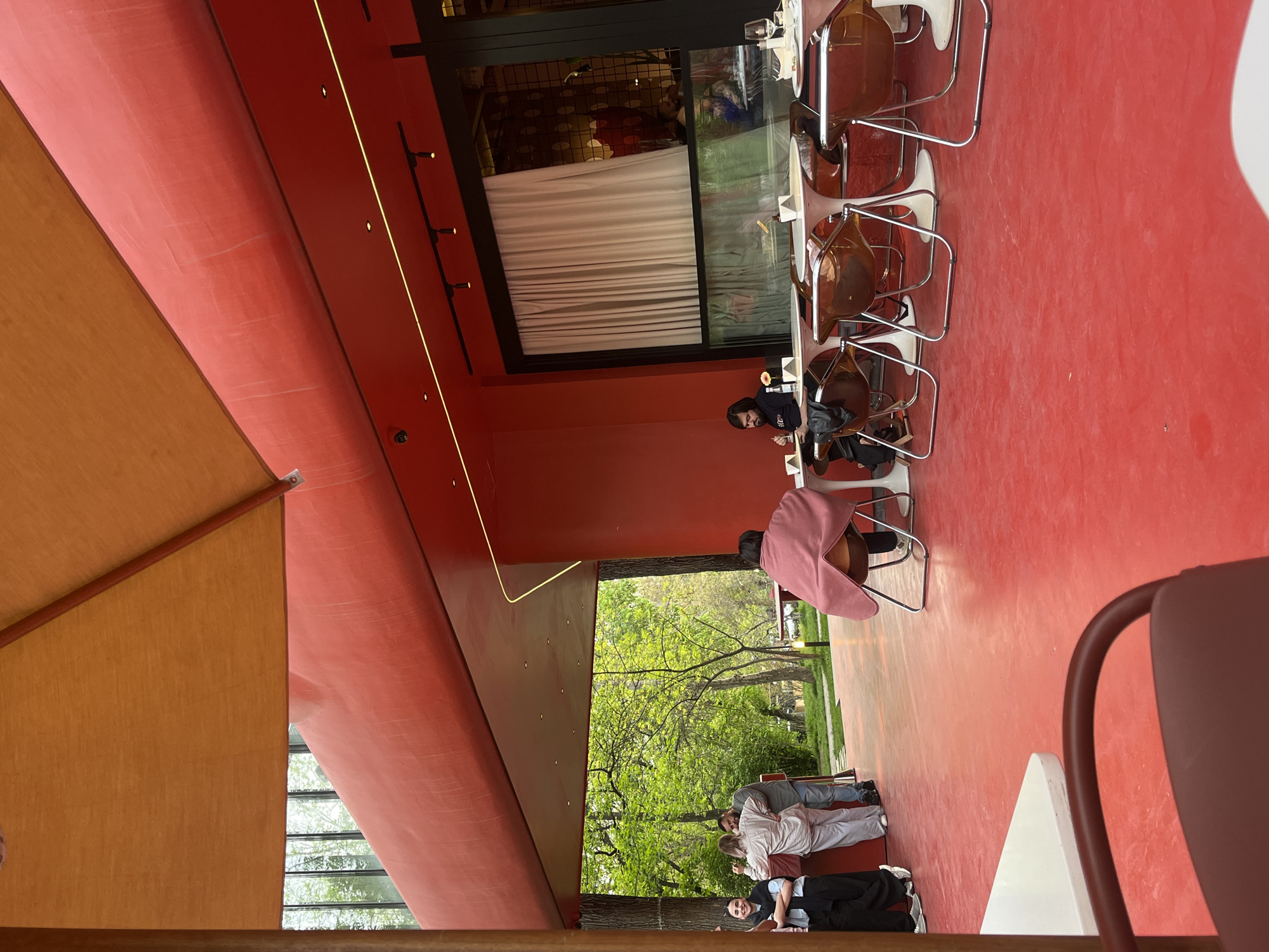 The image captures a scene inside a restaurant or cafe, likely in Yerevan, Armenia, given the architectural style. The dominant color inside is red, covering the floor and ceiling. There is a long table with multiple chairs, occupied by several people. One person is seated alone, seemingly engaged with their meal or device. A couple of people are sitting nearby, and further in the background, a group of people is standing and interacting, possibly waiting for a table or just socializing. A window offers a view of a green space with trees, suggesting a park or garden outside. The lighting is a mix of natural light from the window and artificial light inside.