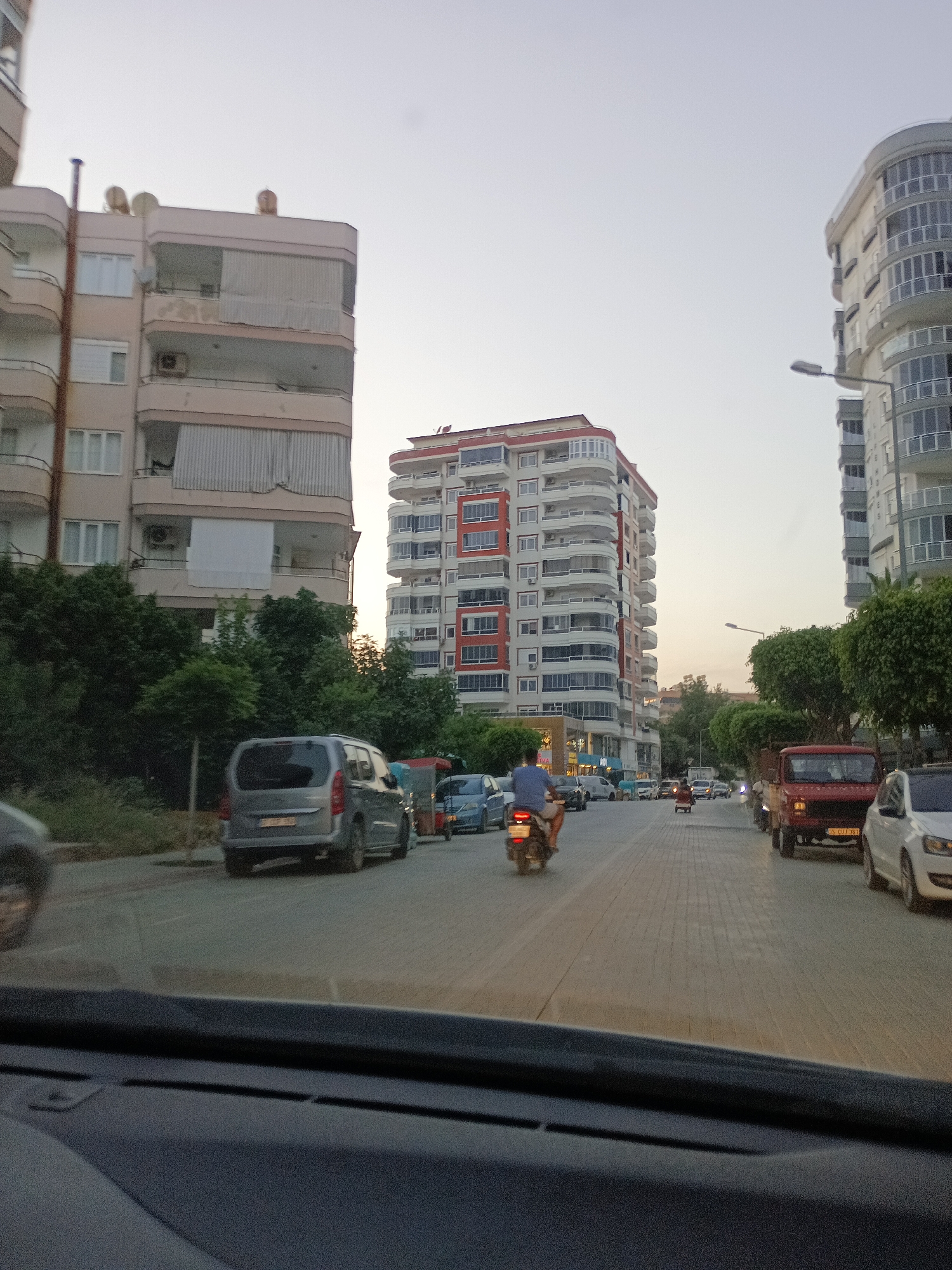 This is a street-level, horizontal, eye-level, full shot of a street in Alanya, Turkey, with buildings lining both sides. The sky is overcast and light orange, suggesting either dawn or dusk. A person rides a motorbike in the left lane. The street is paved with cobblestones. On the left side of the street are a row of trees and a multi-story apartment building. On the right side of the street are more trees, a red jeep, and a white car. In the background, there are more apartment buildings.