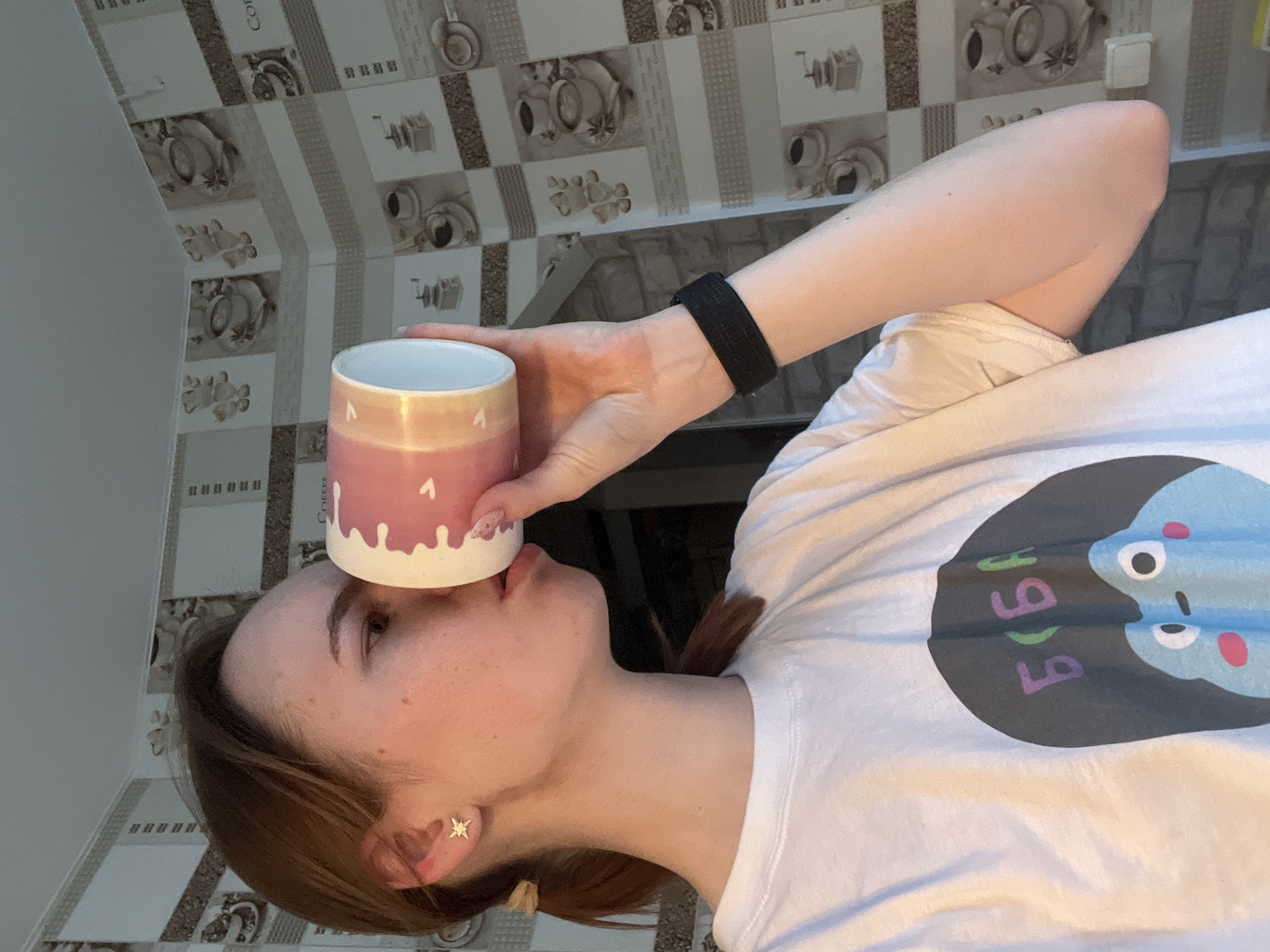 A fair-skinned young woman with brown hair is positioned in front of a patterned wall, likely a kitchen backsplash featuring images of coffee cups and grinders. She is wearing a white t-shirt with a blue and gray cartoon character print. A black wristband is visible on her left wrist. The woman is holding a pink and iridescent mug with white drip designs on its rim, pressed against her nose. She has a star-shaped earring on her left ear. The location is possibly Chaikovsky, Russia.