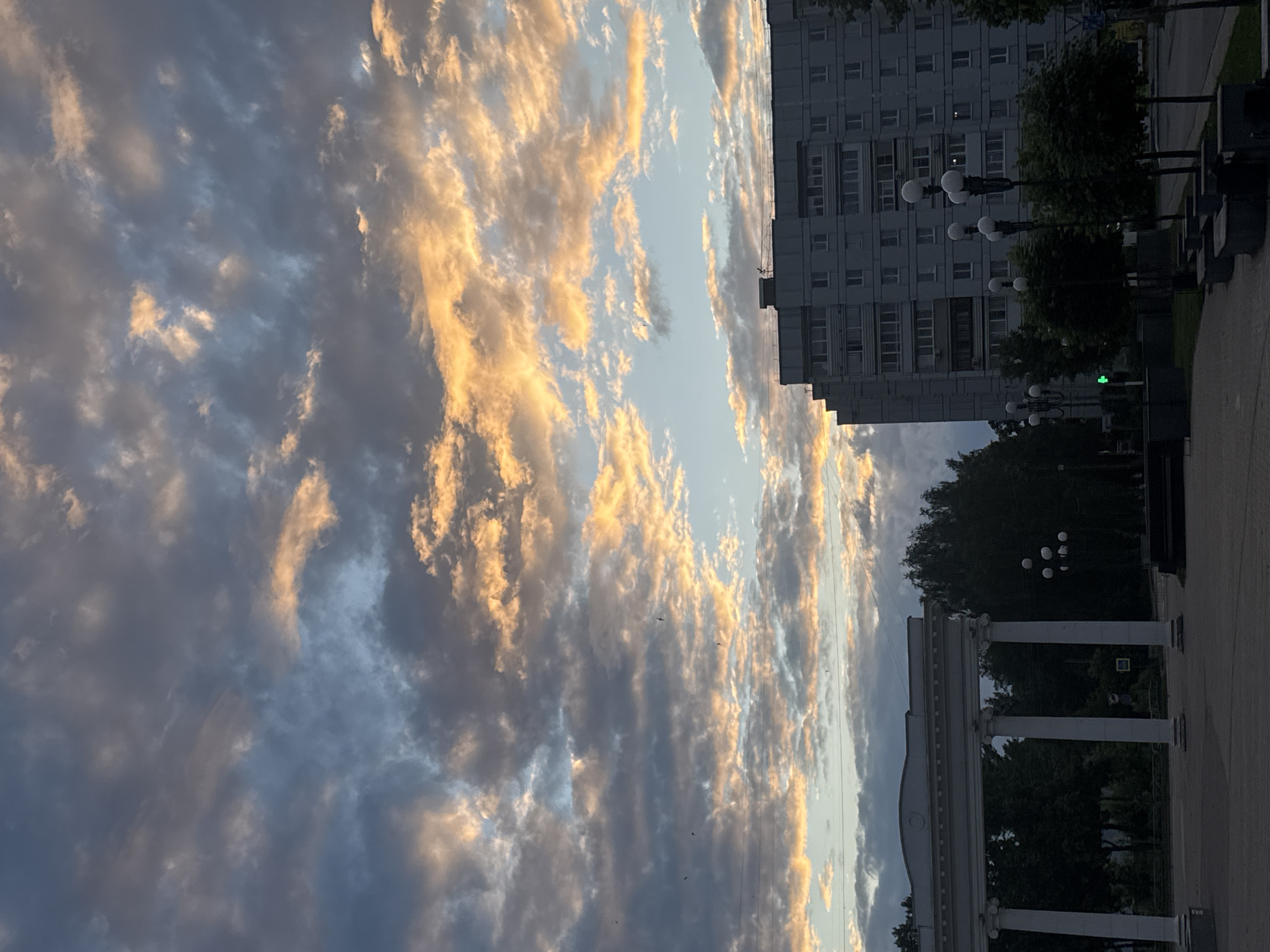 The image depicts an urban scene in Chaikovsky, Russia, dominated by a dramatic sky filled with an array of clouds tinged with the golden and orange hues of sunset or sunrise. Several birds are visible in flight against the colorful clouds. In the foreground, a white classical colonnade stands on the left, partially framing a view of trees and a dark cityscape. To the right, a multi-story apartment building, typical of Soviet-era architecture, rises against the sky. Several streetlights with multiple globes are positioned along a paved area in front of the building and trees. The overall impression is a blend of natural beauty in the sky and the utilitarian aspects of a Russian urban environment.