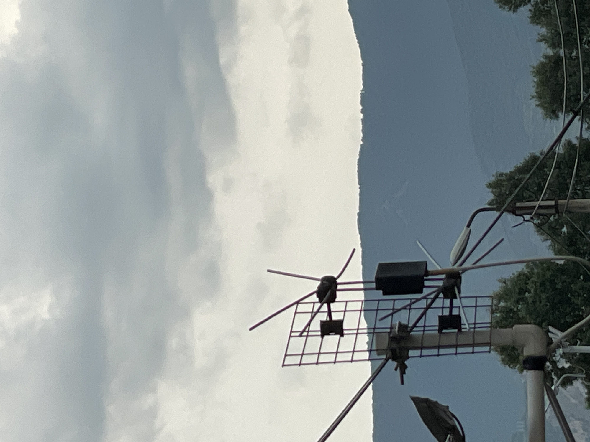 The image shows a close-up of an outdoor television antenna against a cloudy sky and a distant mountain range. The antenna is a multi-element directional type with a grid reflector. There are also some other antennas and wires visible in the foreground, along with some green trees and foliage. The overall scene is somewhat overcast, suggesting it might be a cloudy day. The location is identified as Tbilisi, Georgia.