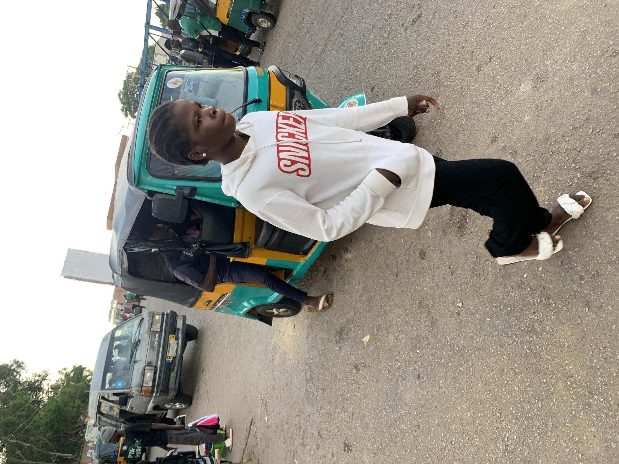 A young Black person wearing a white hoodie with red lettering that says "SNICKERS" and black pants is standing on a paved street, leaning slightly to their right. They are wearing white slides on their feet. Beside them is a green and yellow motorized tricycle, commonly known as a "Keke Napep" or "Marwa." The tricycle appears to be a passenger vehicle. In the background, other vehicles, including a gray minivan with "TOYOTA" written on it and what looks like another tricycle, are visible, suggesting a street market or a transportation hub. The lighting indicates it is daytime, likely late afternoon given the warm tones. The overall scene depicts a bustling urban environment in Jos, Nigeria, likely a place where these tricycles are used for public transportation.