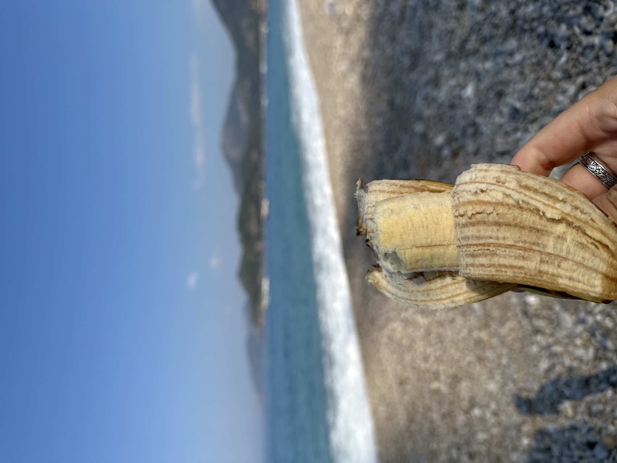 A hand is holding a partially peeled banana in the foreground, with a rocky beach, turquoise water, and distant hills under a clear blue sky in the background. The scene is likely on a beach in Fethiye, Turkey, given the coastal landscape and clear weather.