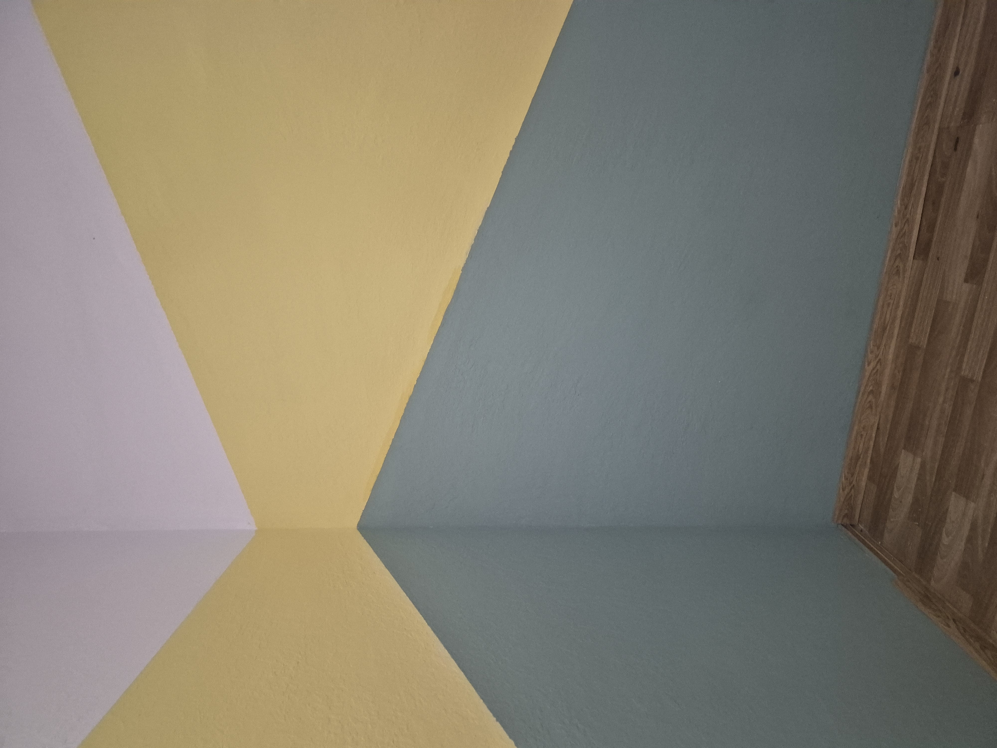 The image shows an interior corner of a room with walls painted in three distinct colors: white at the top, yellow in the middle, and a muted blue-green at the bottom. The paint lines create a geometric, diagonal pattern. The floor is covered with laminate flooring, featuring a wood grain pattern with various shades of brown. The lighting appears to be from an overhead source, casting subtle shadows. There are no people or specific objects of interest visible, making the scene a study in the architectural and color design of the space. The specific location in Yerevan, Armenia cannot be determined from the visual information provided.