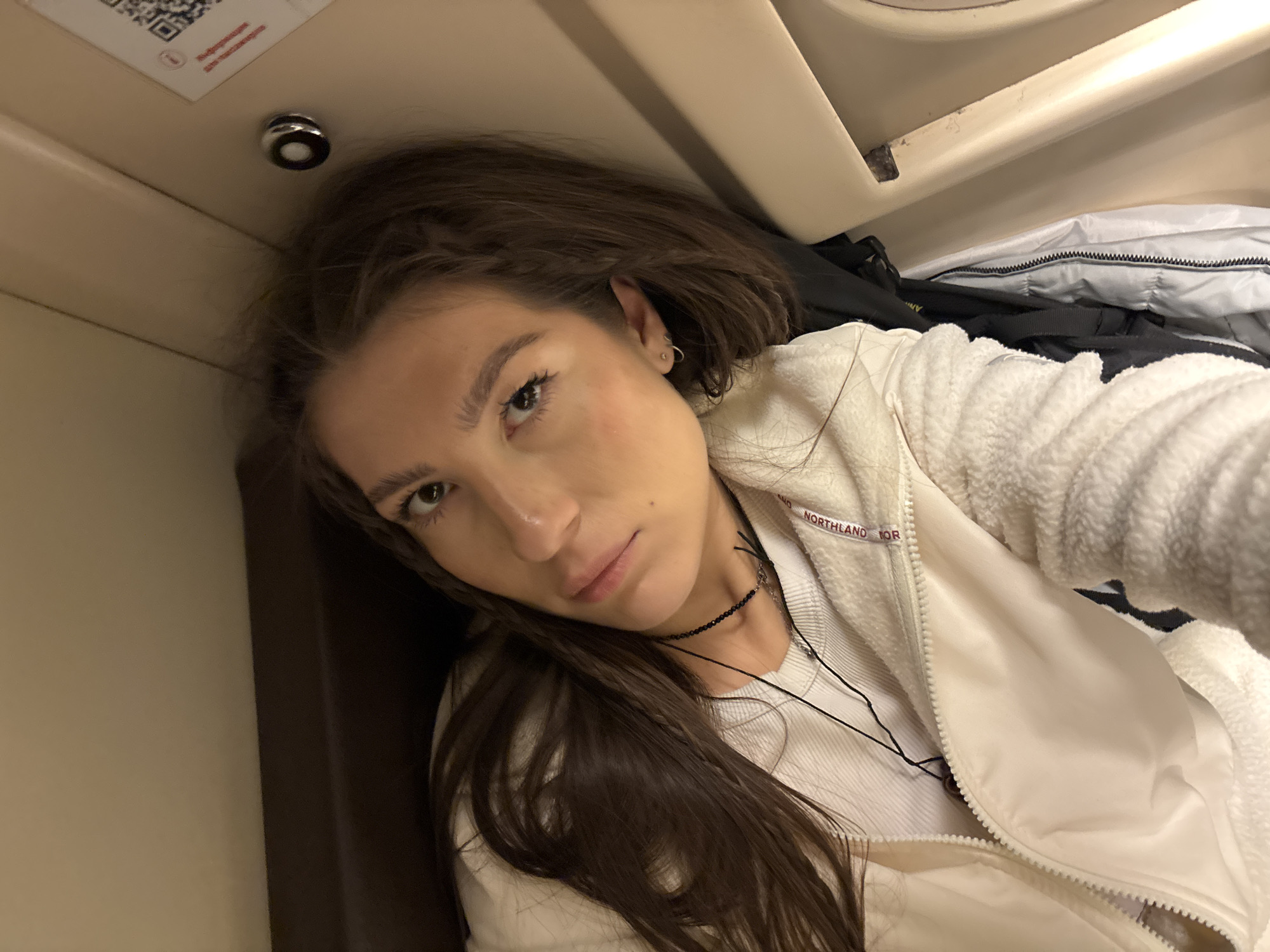 A young woman with long brown hair, wearing a white fleece jacket and a black beaded necklace, is pictured from a close-up, tilted angle. She is indoors, likely in a compartment on a train or similar transportation, as evidenced by the overhead storage and brown-paneled walls. The environment appears confined. The location is potentially near Шемордан, Russia. There is a QR code visible in the upper left corner.
