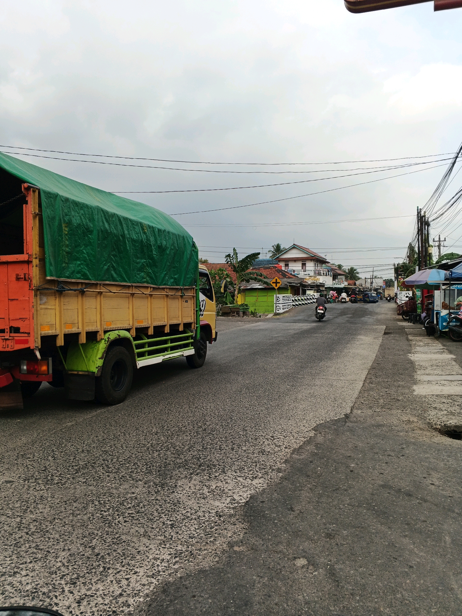 A medium shot captures a truck on a paved road in what appears to be a commercial or residential area under an overcast sky. The truck, painted orange at the rear and yellow with a green tarp cover on its cargo bed, is in the left foreground and is moving away from the viewer and slightly to the right. Its wheels are black, and the chassis has green and white detailing.
The road is asphalt with visible wear and tear. To the right of the truck, a sidewalk with some shops and stalls is visible. One stall has a blue and white awning and appears to be selling food, with the word "goreng" (fried) partially visible. Other stalls are to its left and right, with various awnings and signage. Several motorcycles and a few cars are on the road, some parked and some in motion.
Buildings line the street, featuring tiled roofs in red and orange, and white walls. Some of these buildings appear to be shops, with signs like "MATAMARI" visible. A yellow road sign with a black cross is also present. Utility poles with numerous wires are scattered along the street, indicating it's a populated area. The overall atmosphere is that of a busy street scene on a cloudy day.