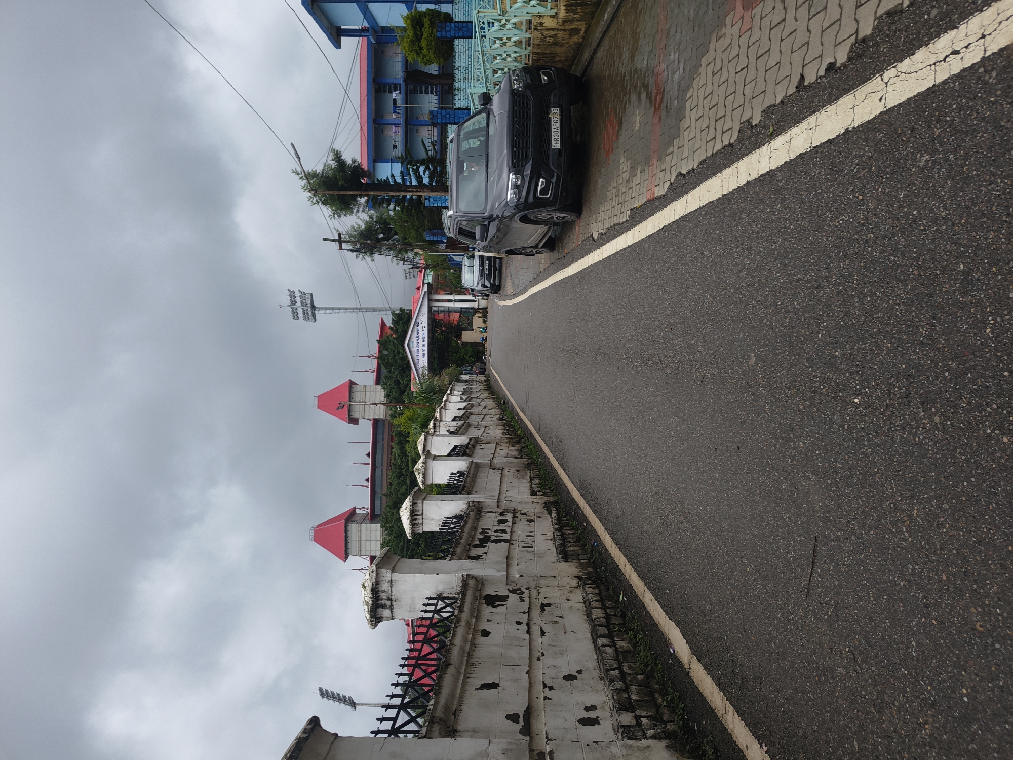 A black Mahindra Scorpio SUV is parked on the right side of a wet, paved road in Dharamshala, India. The SUV has the license plate HR30AF6793. To the left of the road is a white stone wall with black metal fencing. In the background, there are buildings with red roofs, stadium lights, and a cloudy sky. The scene appears to be in a city, possibly near a stadium or public venue, given the architecture and lighting. The road is wet, suggesting it has recently rained or is still raining.