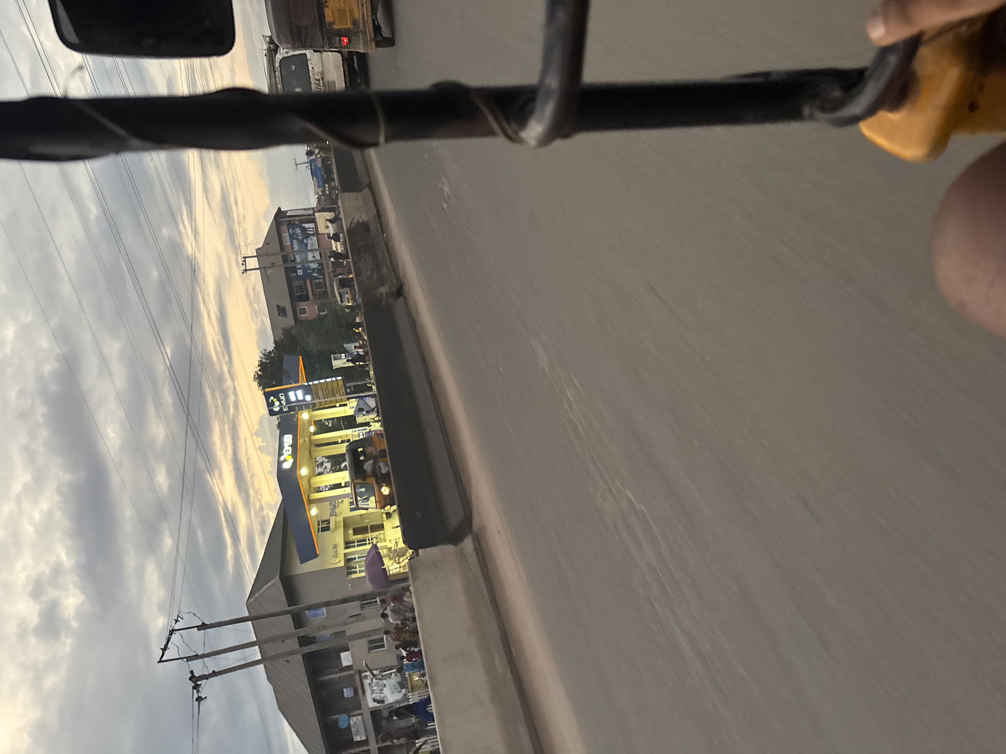 The image captures a view from a moving vehicle, likely a three-wheeled auto-rickshaw, on a street in Onitsha, Nigeria. The scene depicts a bustling roadside with a gas station, identified by its ENJOY branding and bright yellow canopy, as a prominent landmark. Adjacent to the gas station are buildings, one of which appears to be a multi-story structure with people gathered in front of it. Several vehicles, including a white van and another auto-rickshaw, are visible on the road. The sky is cloudy with a hint of sunset colors. The foreground of the image is dominated by the interior of the vehicle, with a black metal pole and a yellow handle. The perspective suggests the image was taken during the day, possibly late afternoon.