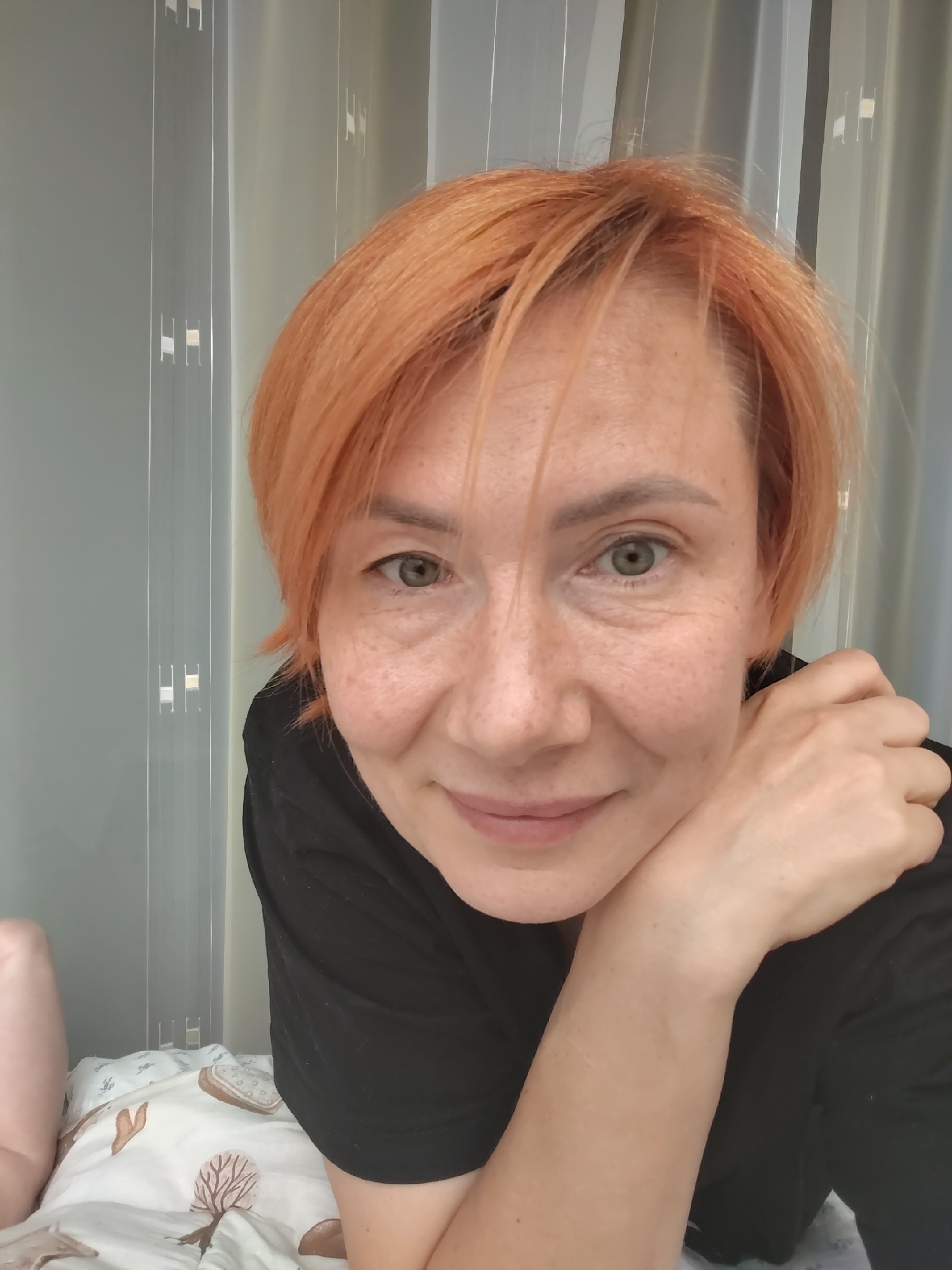 A close-up, head-and-shoulders selfie shows a woman with short, reddish-orange hair and fair skin with freckles. She is wearing a black t-shirt and resting her chin on her hand, which is propped on her arm. Her gaze is directed towards the camera, and she has a slight smile. The background consists of sheer, vertically pleated curtains, some appearing to be translucent and others opaque, suggesting an indoor setting. The lighting is soft and diffused. The image quality is good, with clear focus on the woman's face. No location information is visible in the image itself; therefore, no specific location in Konakovo, Russia can be determined from the provided image.