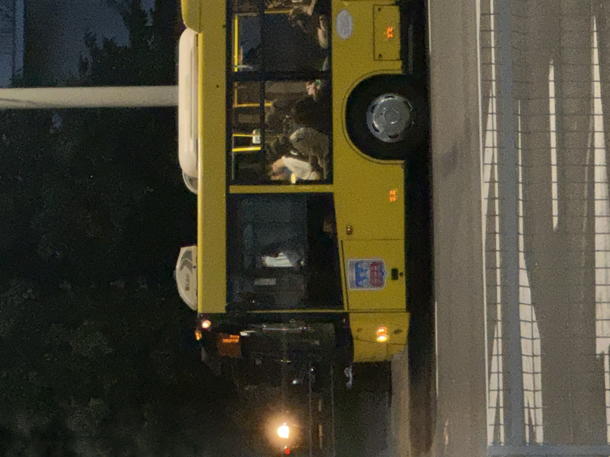 An exterior medium shot captures a yellow public bus at dusk on an urban street, with a pedestrian crossing visible in the foreground. The bus, illuminated by its headlights and interior lights, is positioned at an angle, showing its side profile. Passengers are visible through the tinted windows, seated inside the bus. The bus features a circular emblem on its side, and the "KAYY" logo is visible on the roof. The environment is dark, with trees in the background and a streetlamp glowing on the left side of the frame. The pedestrian crossing is marked with white stripes on asphalt.
