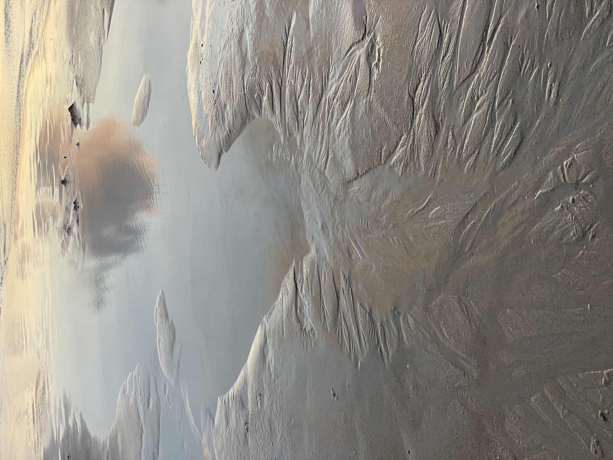 The image shows a close-up view of wet sand on a beach with receding tide lines, creating intricate patterns. Small puddles of water reflect the sky, including a pinkish cloud. There are some small mounds of sand in the water. The scene is devoid of any people or specific structures. The lighting suggests it may be during dawn or dusk. The location is identified as Rawai, Thailand.