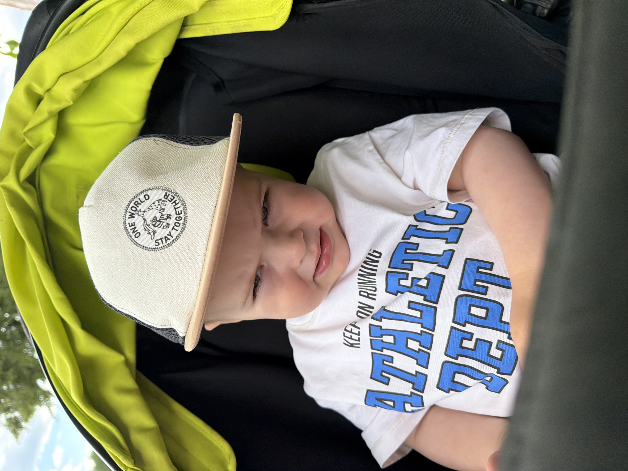 A close-up, eye-level shot shows a Caucasian baby, appearing to be between one and two years old, sitting in a stroller. The baby is wearing a white t-shirt with large blue lettering that reads "ATHLETIC DEPT." and a cream-colored baseball cap with a black mesh back. The cap has a circular logo with the words "ONE WORLD STAY TOGETHER" and images of a giraffe and a zebra. The baby has a slightly grumpy or displeased expression, with eyebrows furrowed and mouth slightly open. The stroller has a bright lime green interior lining and a black exterior. The background is blurred, showing some greenery and a light sky, suggesting an outdoor setting. The image was taken in Votkinsk, Russia.